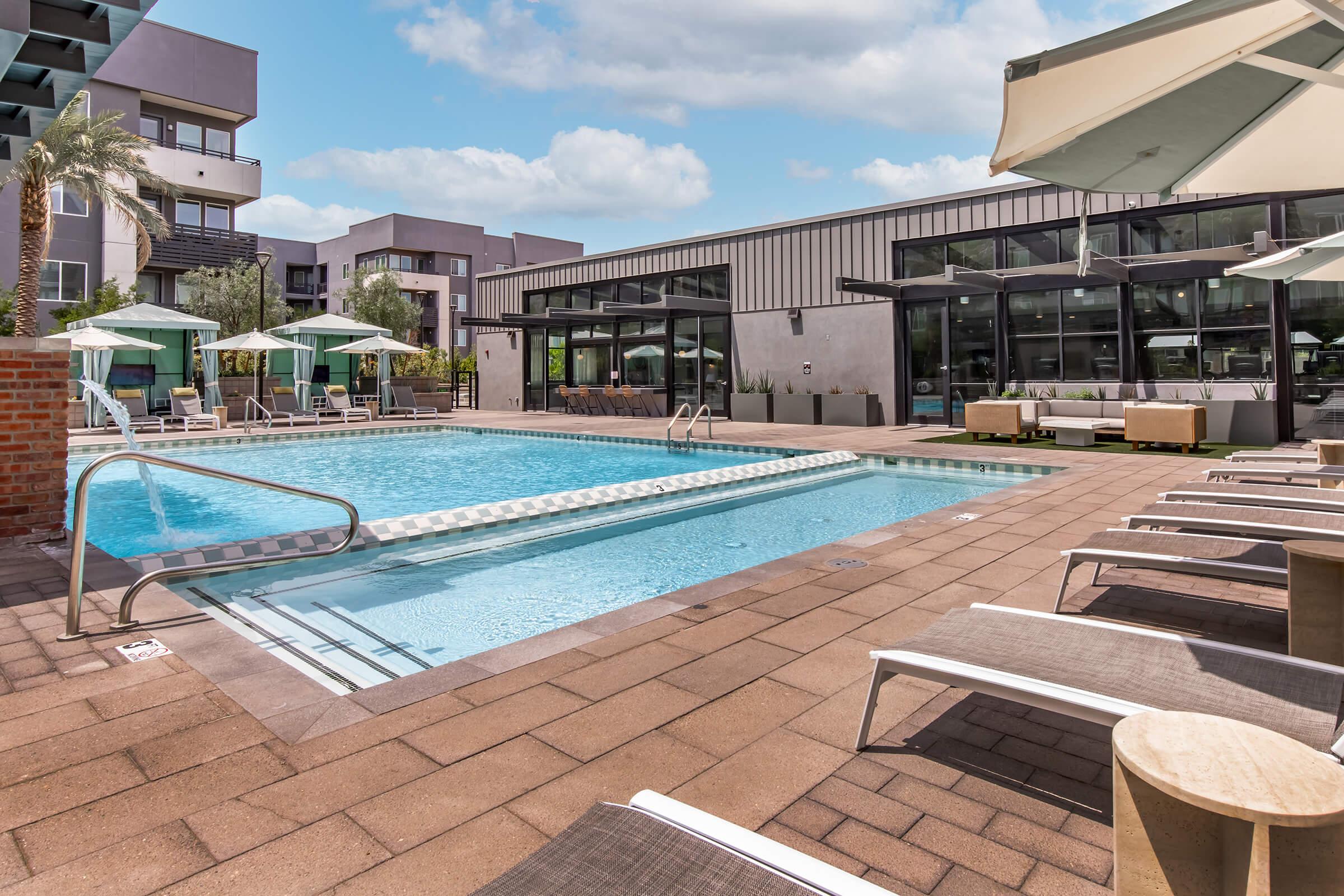 A modern outdoor swimming pool surrounded by lounge chairs and umbrellas, with a contemporary building in the background. The area is landscaped with palm trees, and the sky is partly cloudy, creating a relaxed and inviting atmosphere.