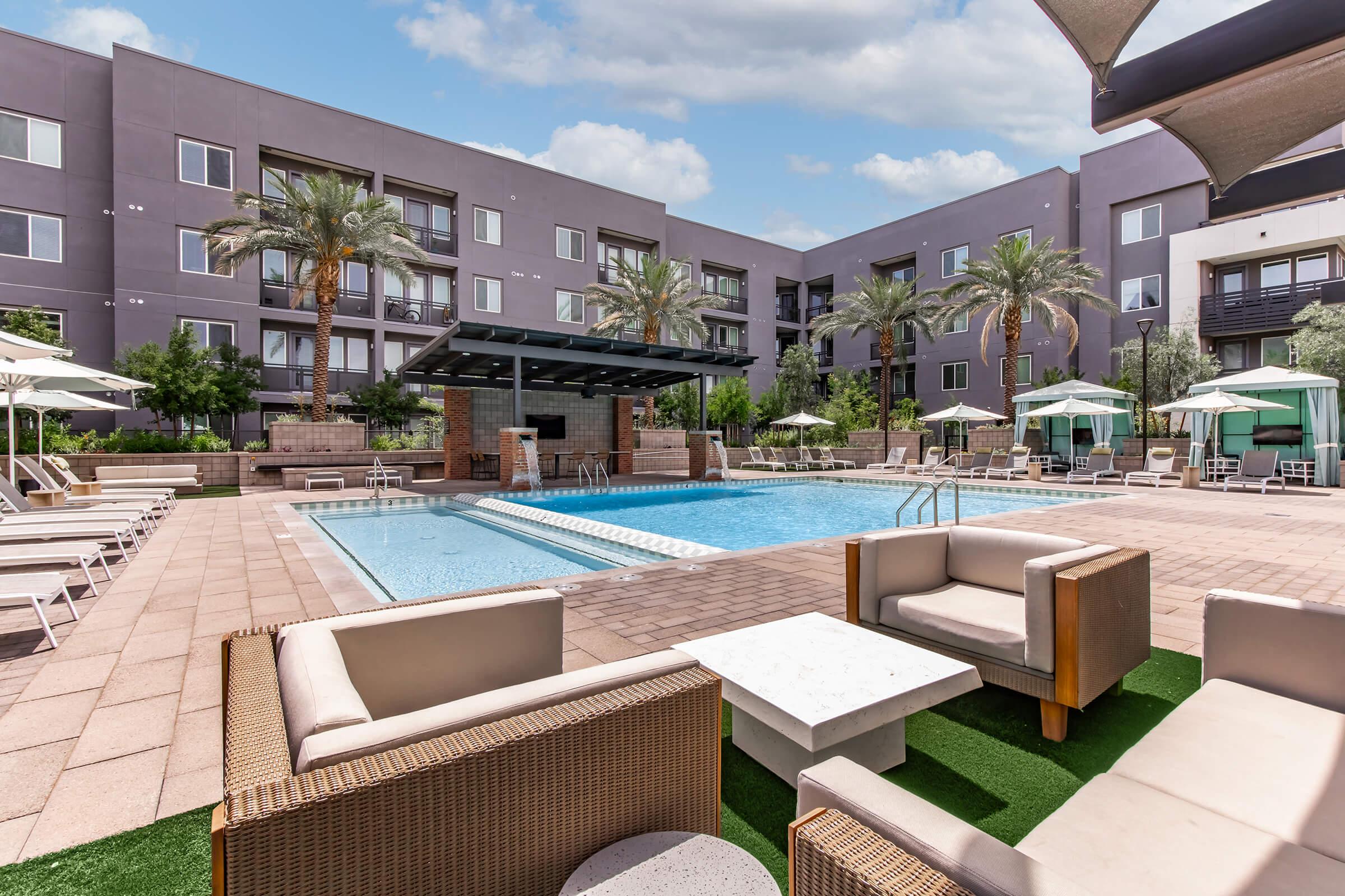 A luxurious outdoor pool area featuring a large swimming pool, lounge chairs, and elegant seating arrangements. Surrounded by palm trees and modern apartment buildings, the space includes shaded areas with umbrellas and a stylish pergola, creating a relaxing ambiance for residents and guests.