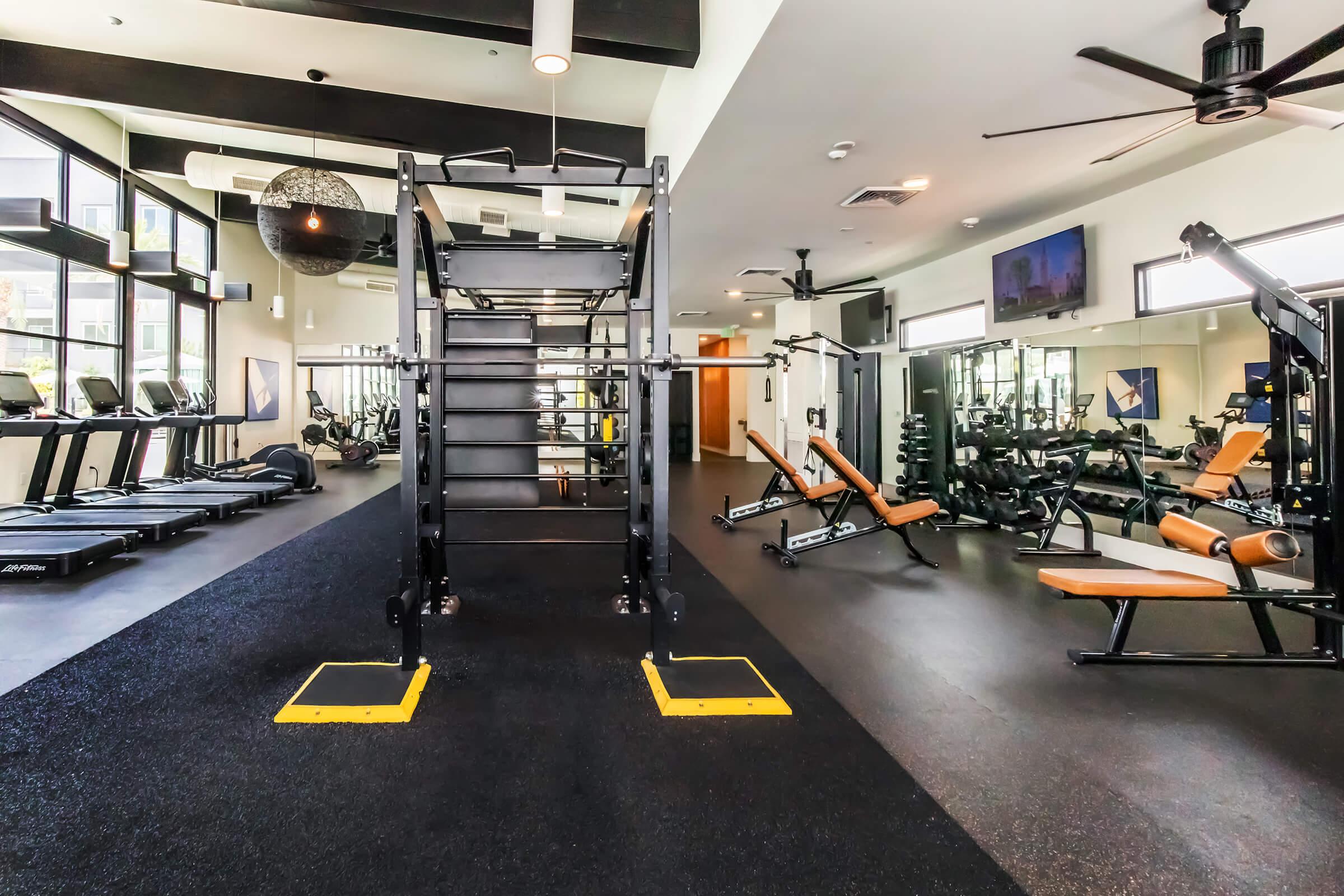 A modern gym interior featuring various fitness equipment. The space includes treadmills, weight benches, dumbbells, and a multi-functional training rig. Large windows provide natural light, and the floor is covered with dark rubber matting for safety. The environment is designed for strength training and cardio workouts.