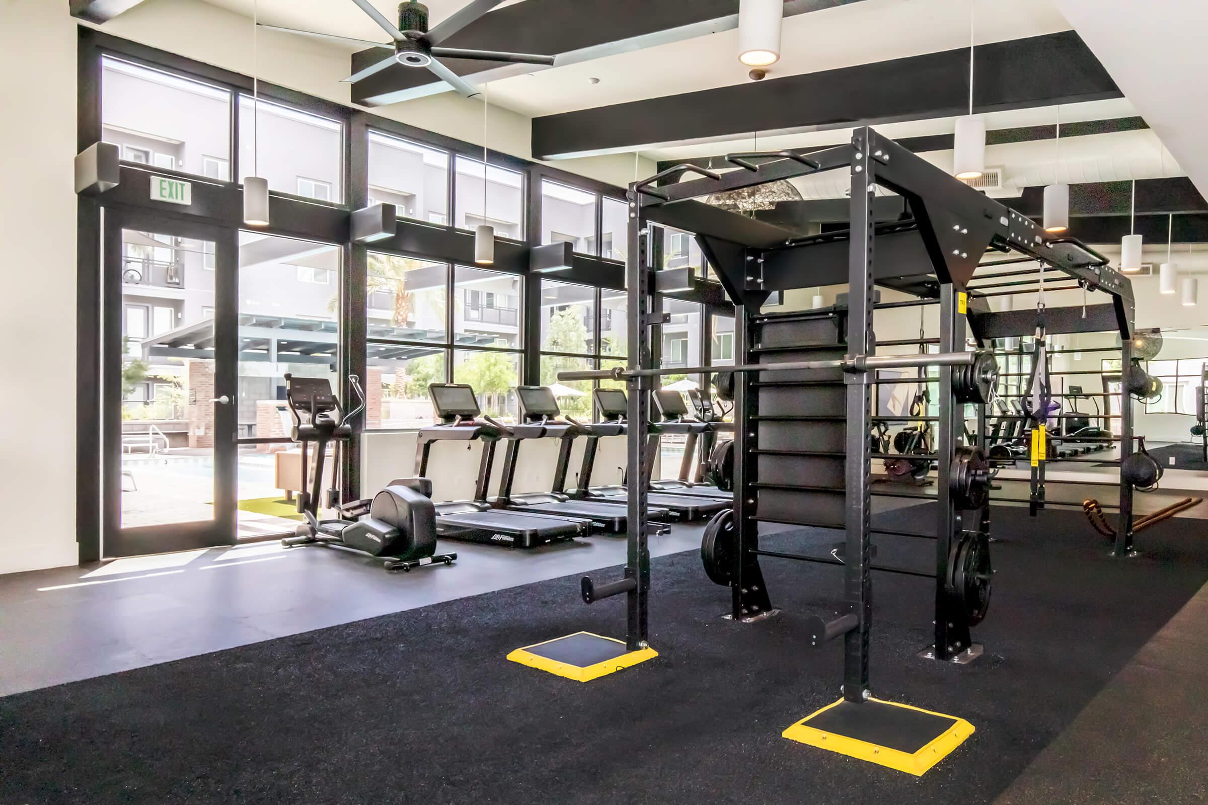 Interior of a modern gym featuring several treadmills, a functional training area with weightlifting equipment, and large windows allowing natural light. The black and yellow color scheme is complemented by sleek design elements and a view of an outdoor space, creating an inviting workout environment.