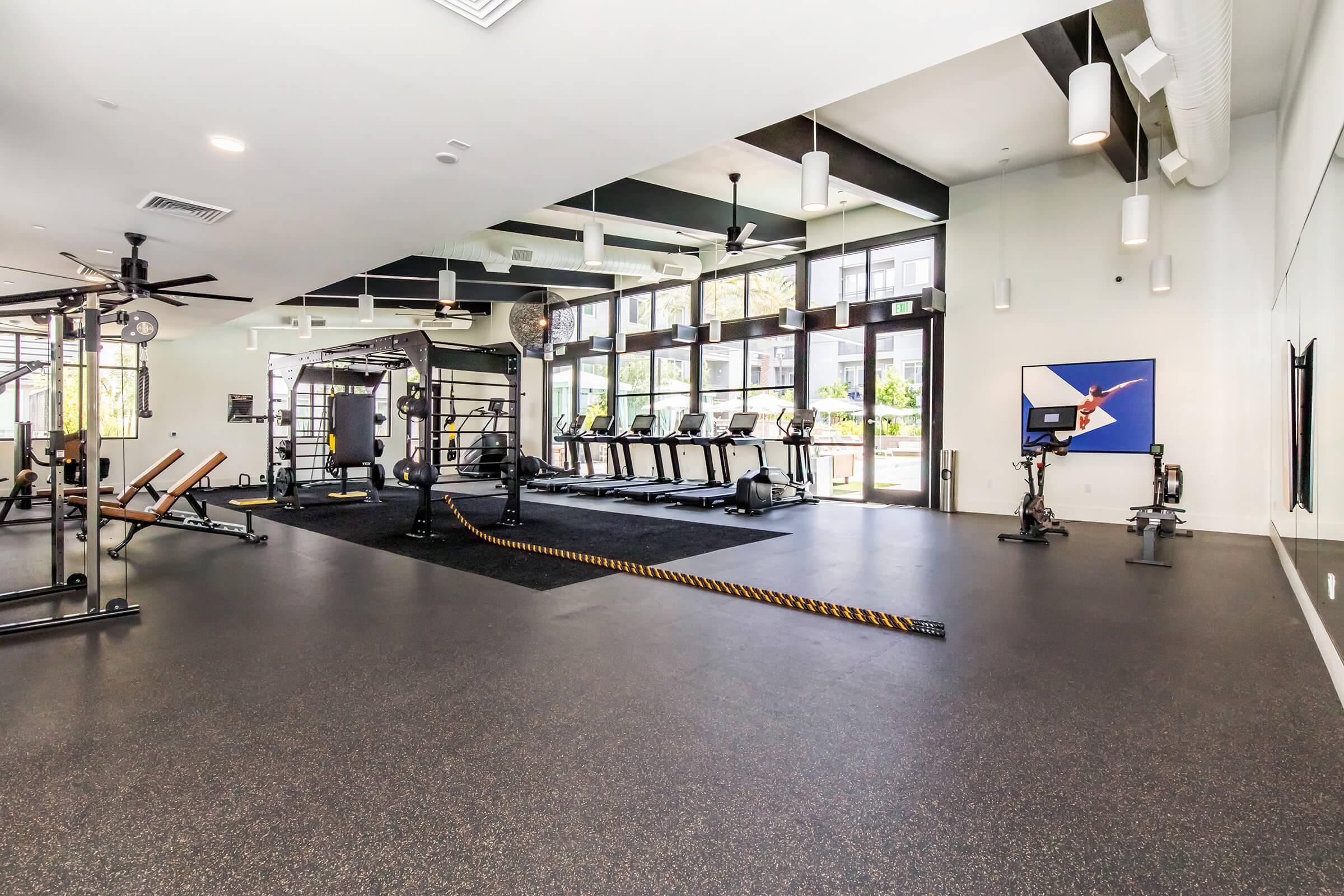 A spacious gym interior featuring various exercise equipment, including treadmills, a rowing machine, and strength training stations. There are large windows letting in natural light, modern lighting fixtures, and a clean, modern aesthetic with dark flooring.