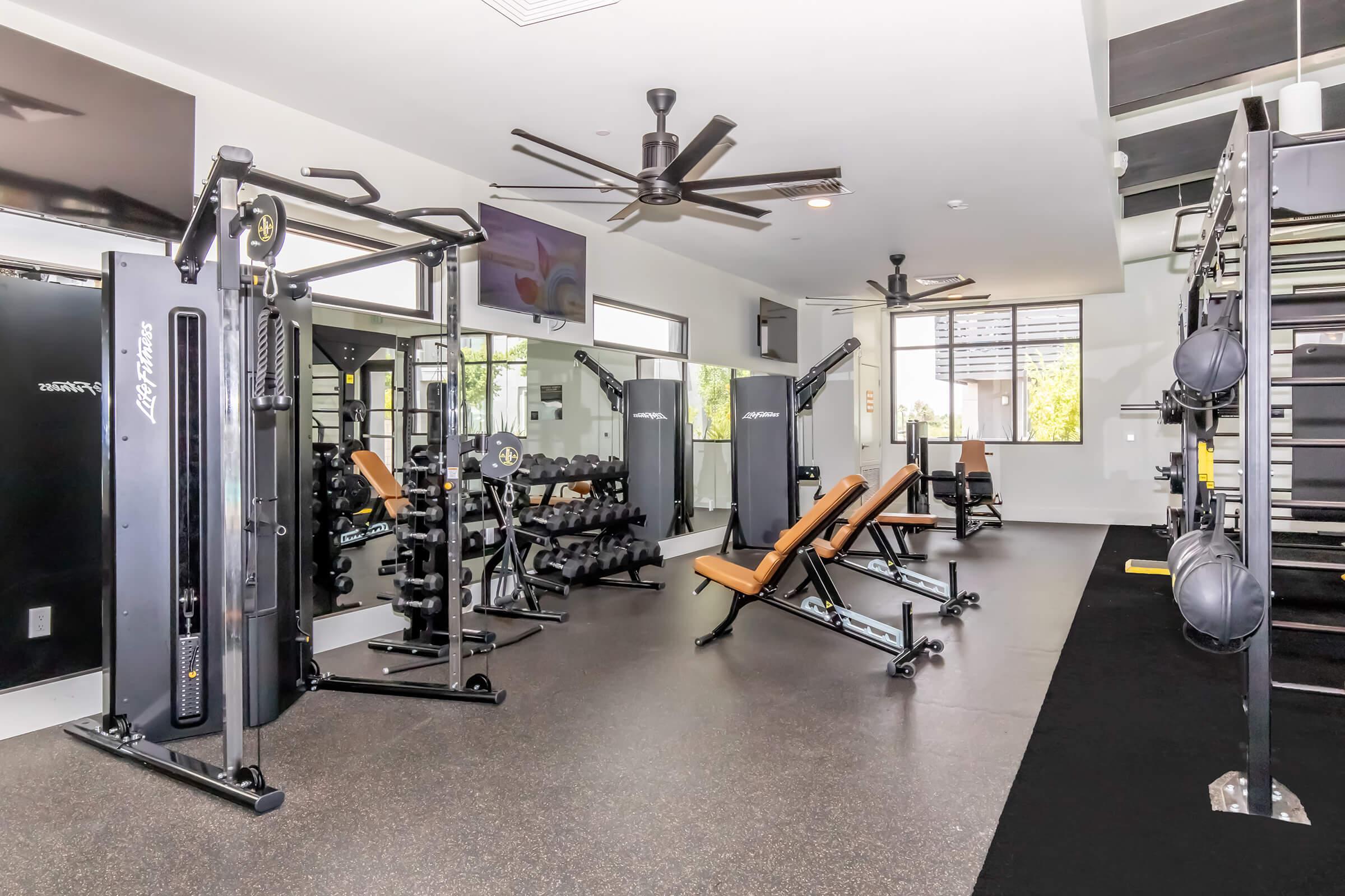 A modern gym space featuring various workout equipment, including weight machines, benches, free weights, and a large mirror. The room is well-lit with ceiling fans and has multiple flat-screen TVs mounted on the walls, creating an inviting atmosphere for fitness activities.