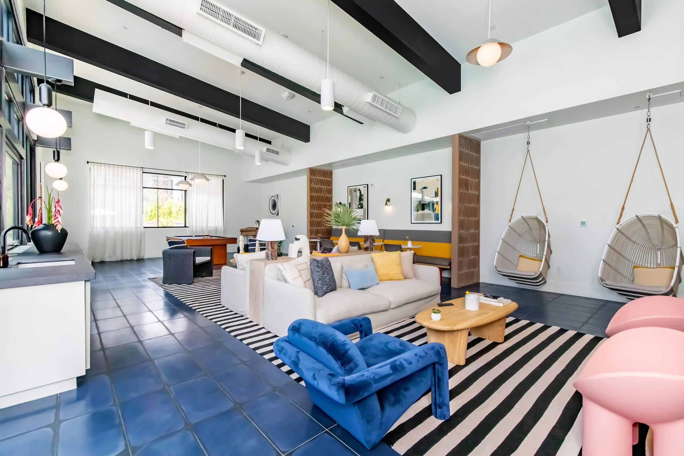 A modern living space featuring a light-colored sofa, blue armchair, and wooden coffee table on a striped rug. Two hanging chairs are present, alongside a pool table. Large windows let in natural light, with plants and decorative artwork enhancing the contemporary decor. The ceiling has exposed beams and industrial lighting.