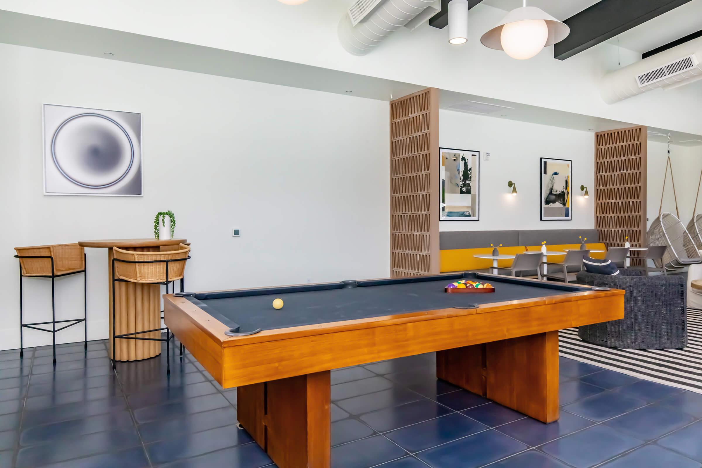 A modern recreational room featuring a billiard table with a cue ball and colored balls arranged. There are two wooden bar stools nearby, along with stylish seating and artwork on the walls. The space is well-lit, showcasing a contemporary design with clean lines and a mix of furnishings.