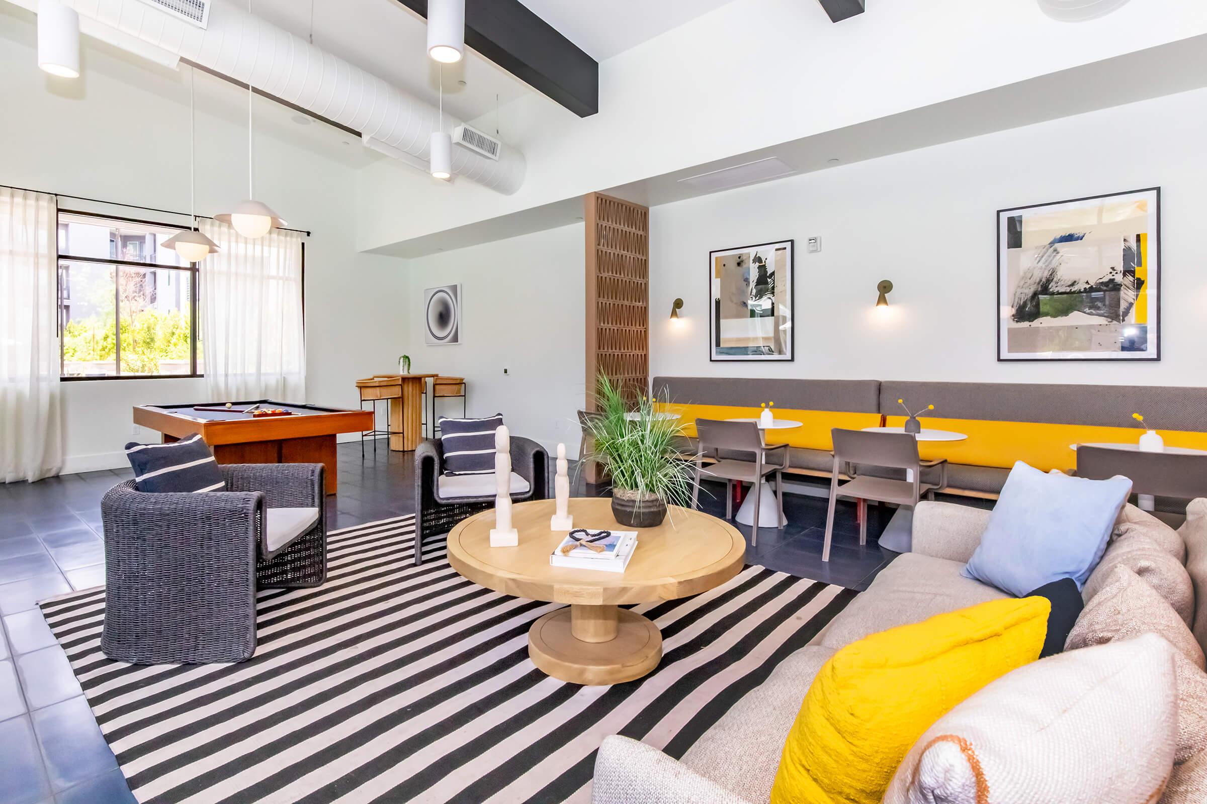 A modern lounge area featuring a striped rug, a round wooden coffee table, and a comfortable sofa with colorful cushions. Large windows allow natural light in, and there are wall art pieces. A pool table is visible in the background, along with dining-style seating arrangements along the wall.