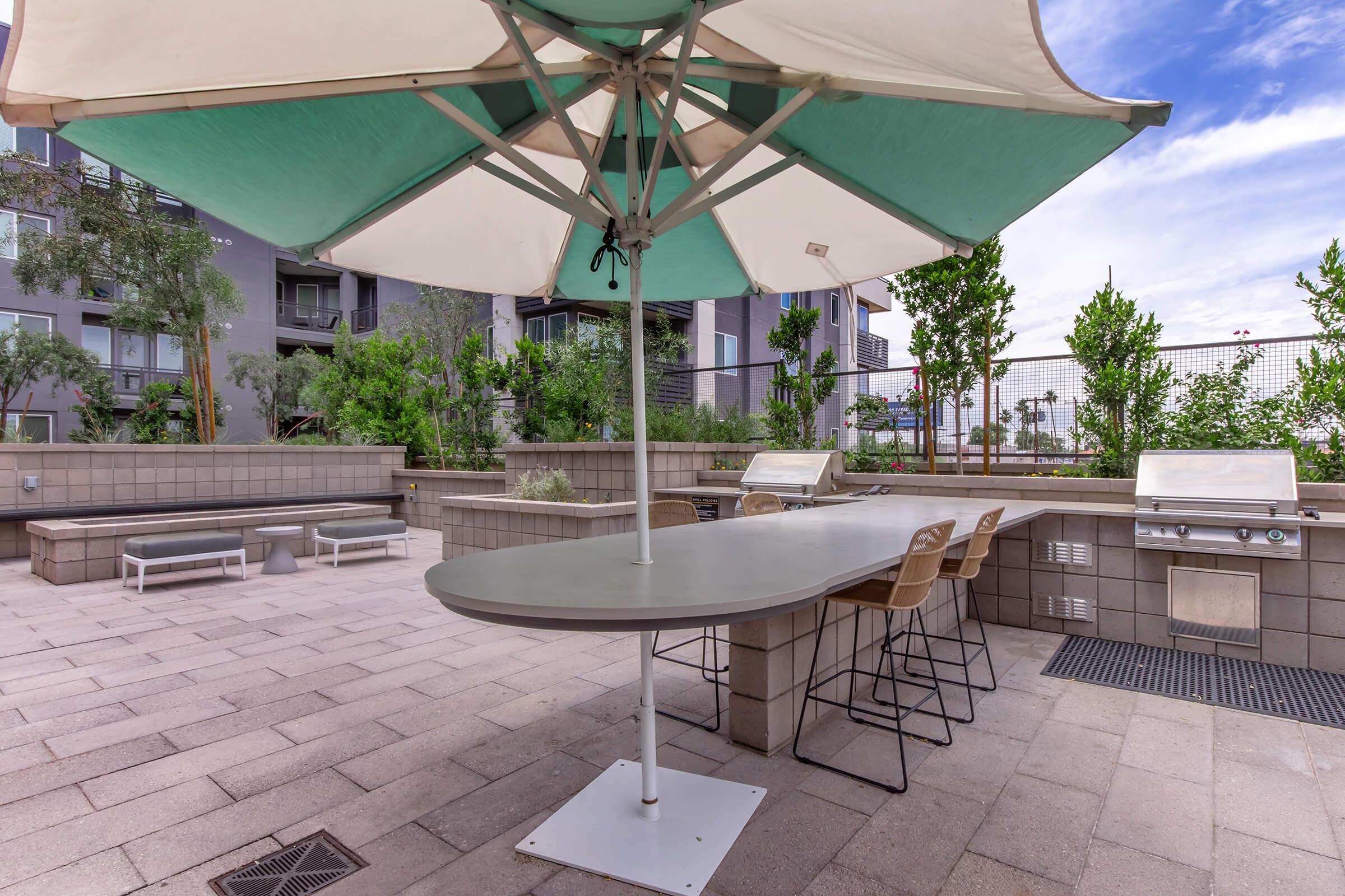 A modern outdoor area featuring a large umbrella over a curved table with bar stools, surrounded by greenery and landscaping. There are built-in grills and seating areas in a neat, contemporary design, set against a backdrop of a residential building. The sky is clear with a few clouds.