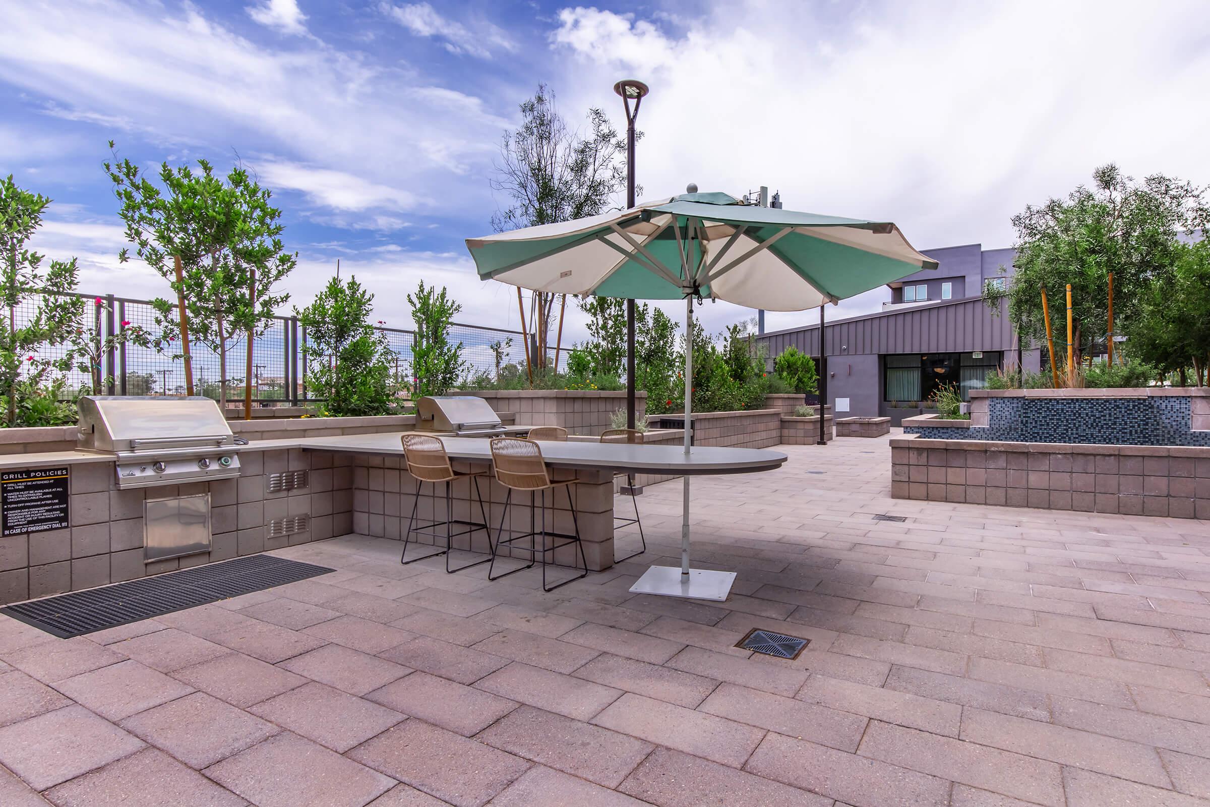 An outdoor space featuring a grill, a stone table with two chairs, and an umbrella. Lush green trees and plants surround the area, with a modern building and a water feature visible in the background. The sky is partly cloudy, creating a pleasant atmosphere.