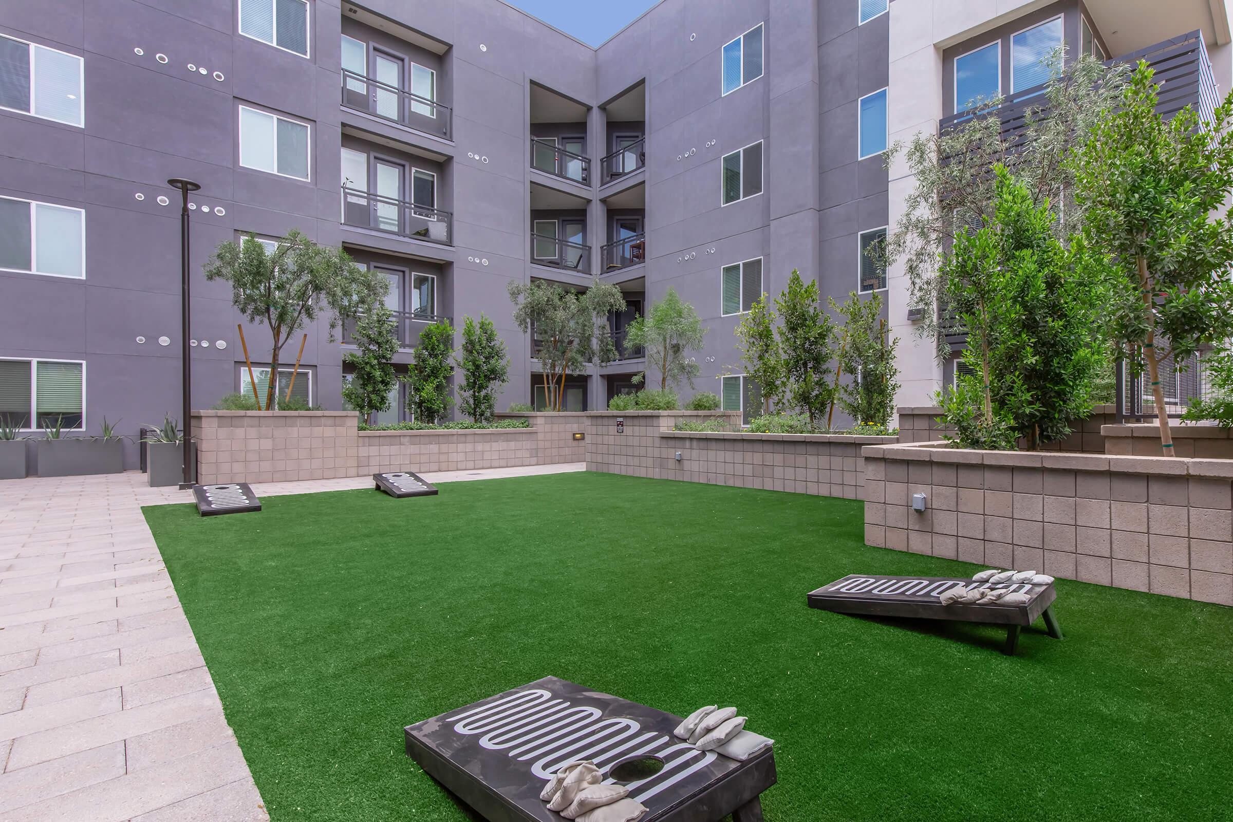View of a landscaped courtyard in a modern apartment complex featuring artificial turf and two cornhole boards. Surrounding the area are potted trees and shrubs, with multi-story buildings and balconies in the background under a clear blue sky.