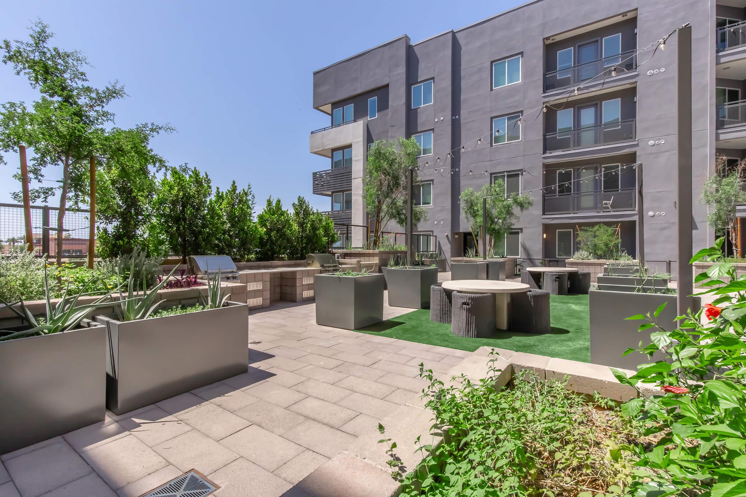 landscaped courtyard with modern seating areas, planters, and artificial grass, surrounded by contemporary apartment buildings under a clear blue sky.