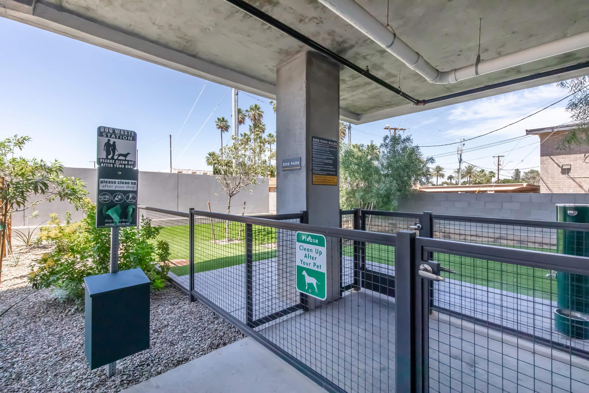 Dog park area featuring green grass, signage for pet waste cleanup, and fenced spaces. The park is situated in an outdoor setting with palm trees and utility poles visible in the background, under a blue sky. The facility promotes a clean and safe environment for pets.