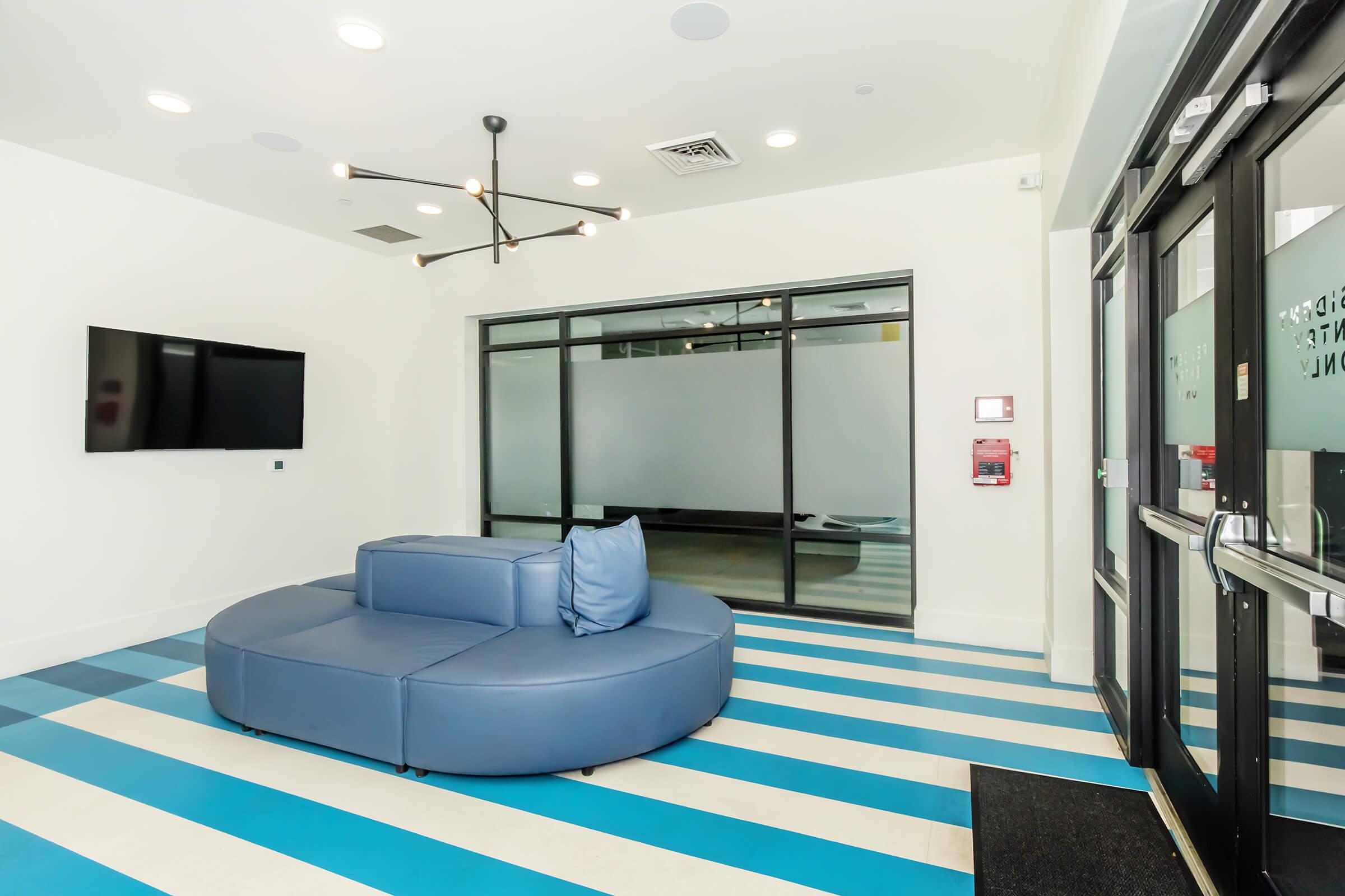 A modern lobby area featuring a large, round blue sofa with a single blue cushion. The floor has alternating blue and white stripes, and a flat-screen TV is mounted on the wall. Glass doors lead to an adjacent space, creating a bright and inviting atmosphere.