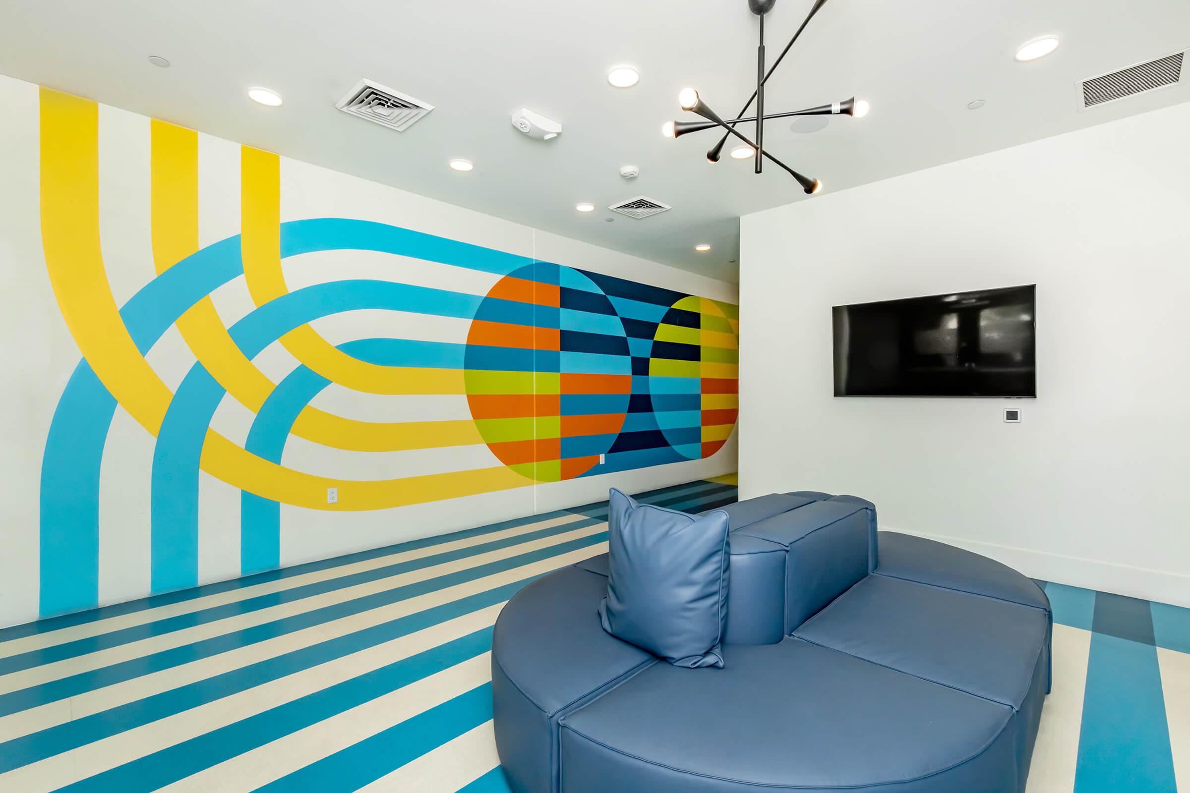 A modern interior with a vibrant mural featuring blue, green, and yellow stripes and circles. The room has a light-colored floor with bold blue and white stripes. A circular blue sofa is in the center, and a black flat-screen TV is mounted on a white wall. The space has bright lighting and a stylish chandelier.