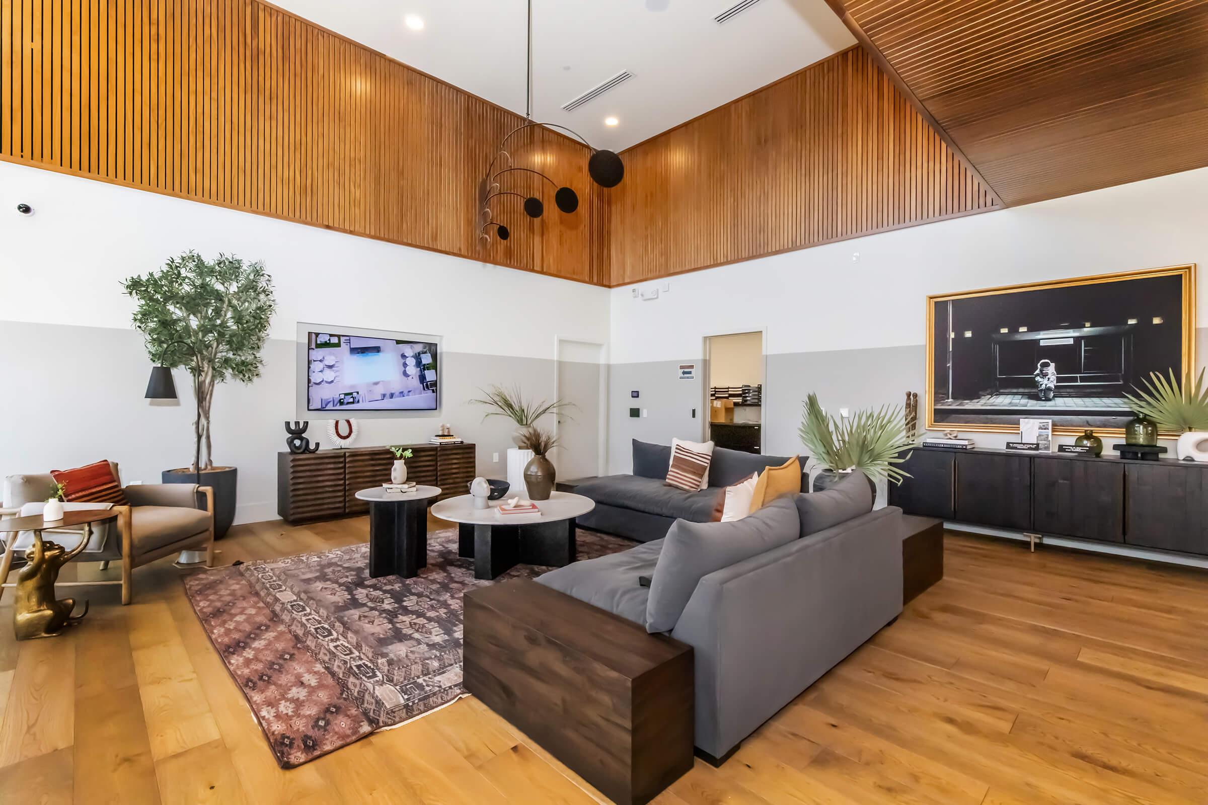 A modern living room featuring a high ceiling with wooden paneling. It includes a gray sectional sofa, a round coffee table, and a decorative rug. A large TV is mounted on the wall, and there are plants and artwork adding style to the space. The floor is wooden, creating a warm atmosphere.