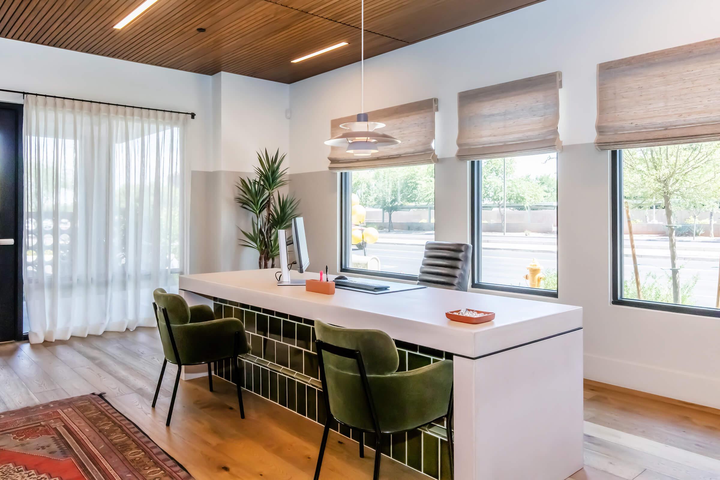 Modern office reception area featuring a sleek white desk with a computer, surrounded by large windows with shades. Two green upholstered chairs are positioned in front of the desk. A potted plant adds a touch of greenery, and a decorative rug is partially visible on the wooden floor. Natural light brightens the space.