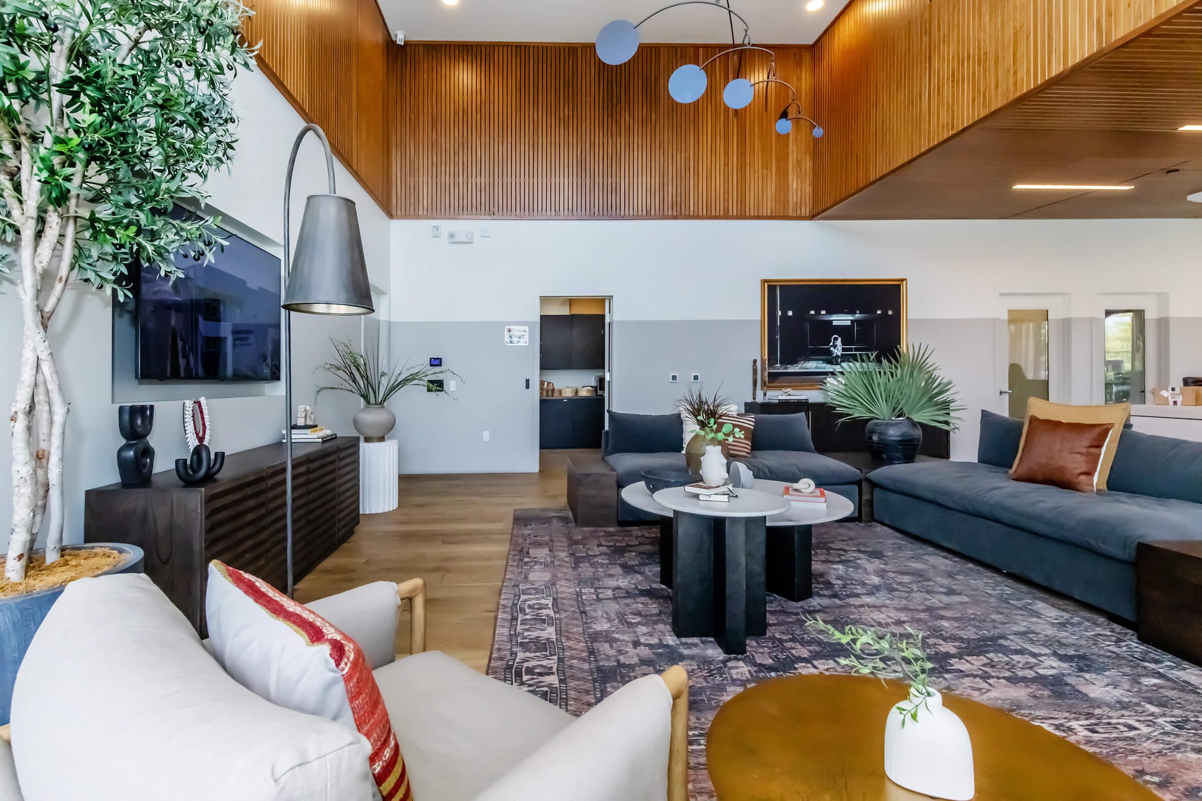 A modern living room featuring a large gray sectional sofa, a round coffee table with a decorative centerpiece, and stylish decor elements. There are potted plants, a floor lamp, and wall art. High ceilings with wood paneling and a contemporary light fixture enhance the spacious feel. A TV is mounted on the wall.