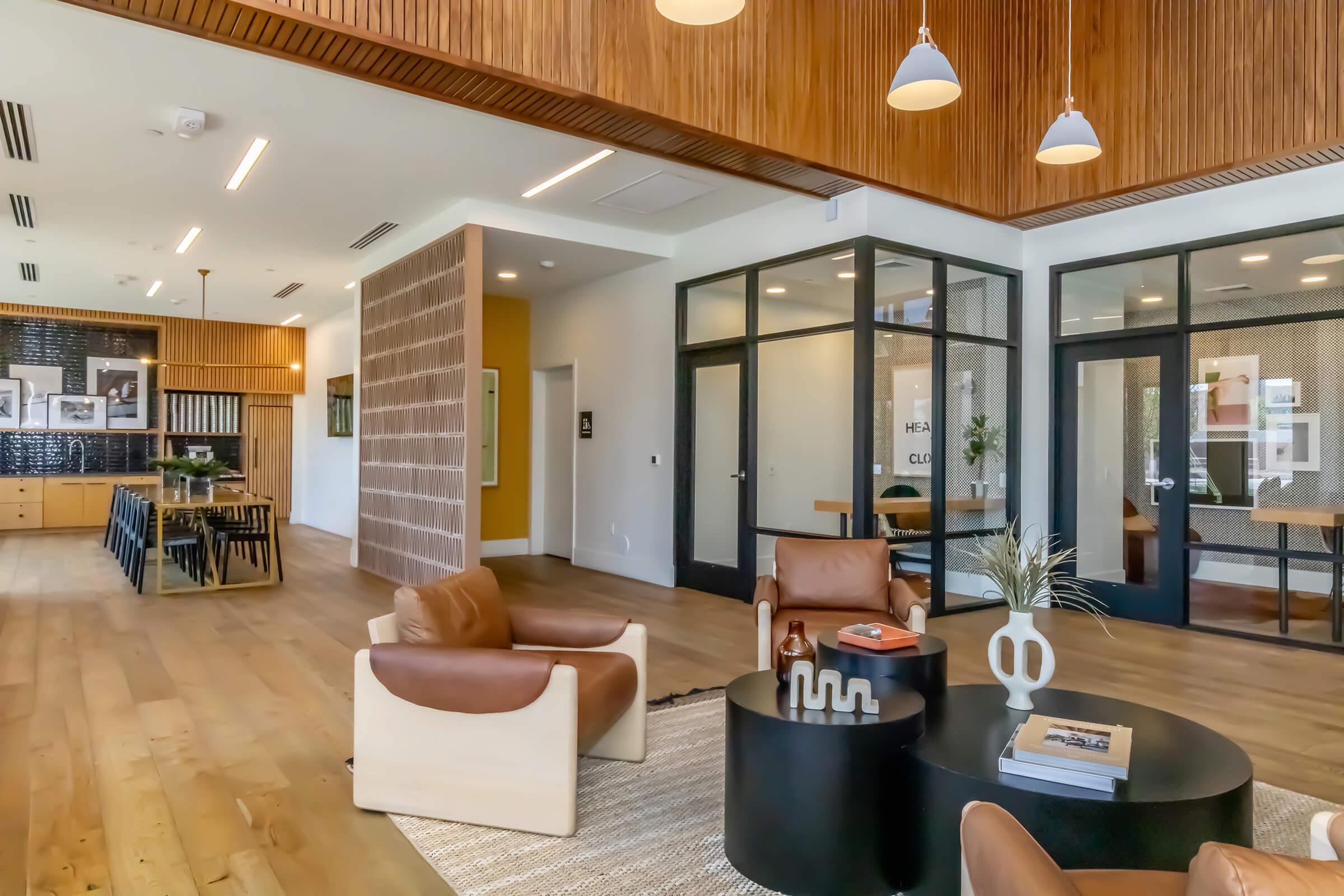 A modern interior space featuring brown leather armchairs, a round coffee table, and a large rug. The room has a wooden ceiling, glass walls showcasing a meeting area, and a dining table in the background. Bright lighting enhances the contemporary design and openness of the area.