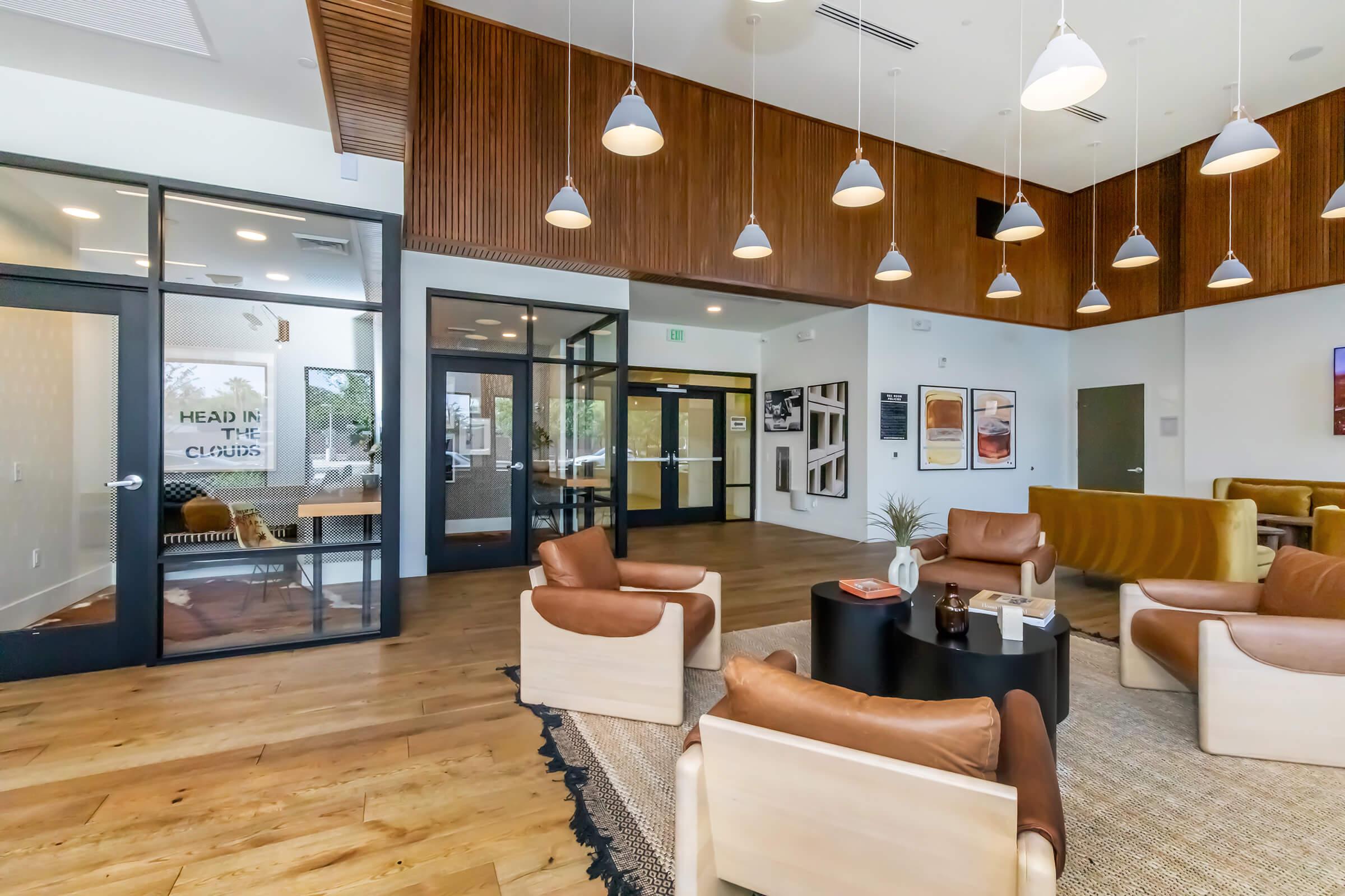 A modern lounge area features leather armchairs, a coffee table, and wood-paneled walls. Pendant lights hang from the ceiling, illuminating the space. Large windows provide natural light, and doors lead to another room with glass walls displaying artwork and seating. The floor is wooden, adding warmth to the contemporary design.