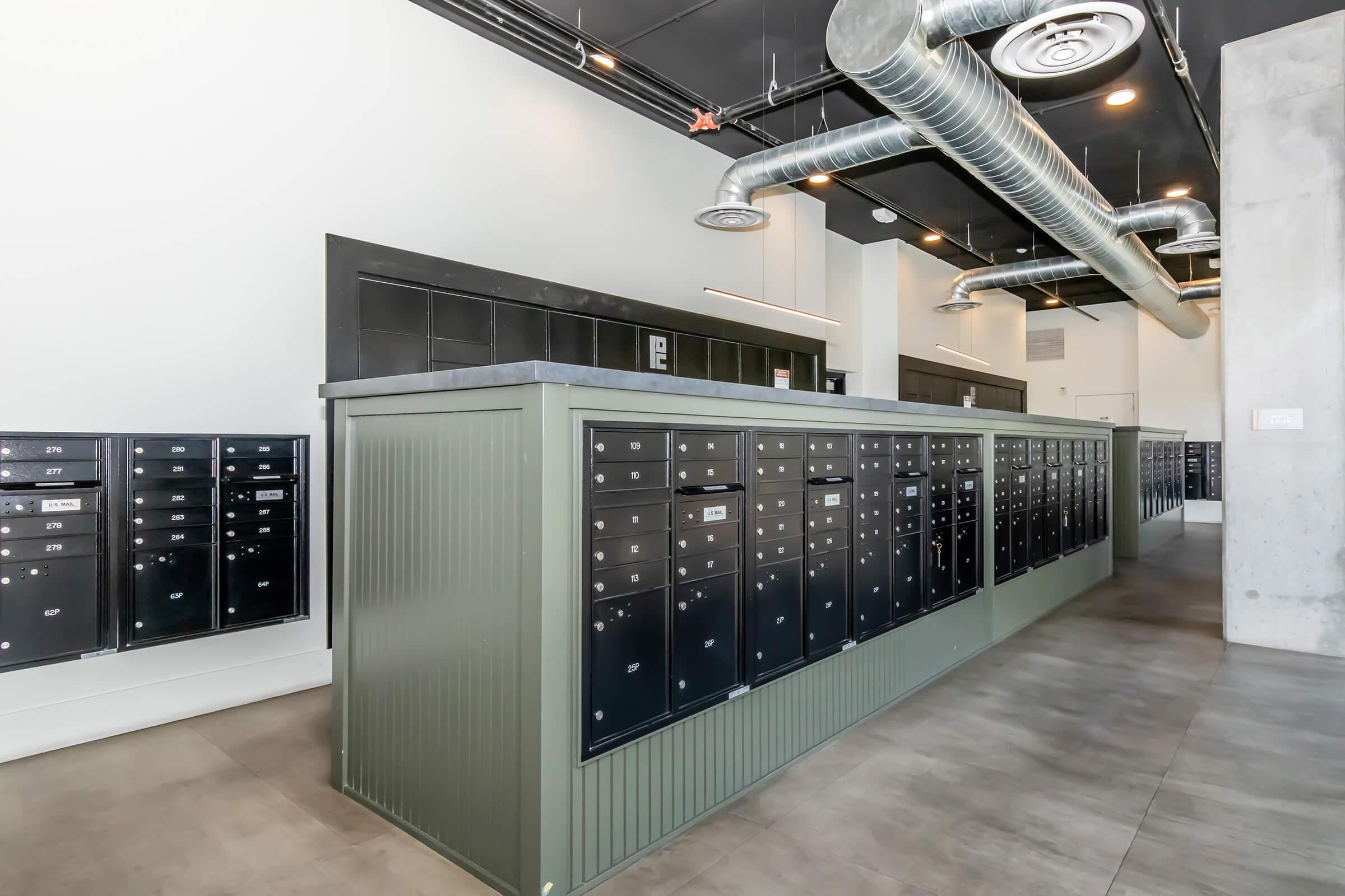 Indoor mailroom featuring sleek, modern mailboxes in a lined arrangement. The space has industrial design elements including exposed ductwork on the ceiling, large windows, and a clean, minimalist aesthetic. The floor is tiled, enhancing the contemporary look.