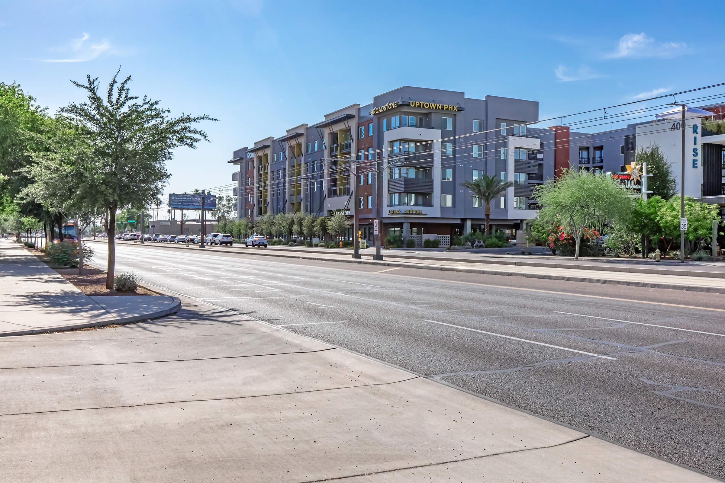A sunny urban street lined with modern apartment buildings and palm trees. The road is empty, showcasing clear blue skies and a few sparse vehicles in the distance. Sidewalks are visible alongside the street, enhancing the suburban feel of the scene.