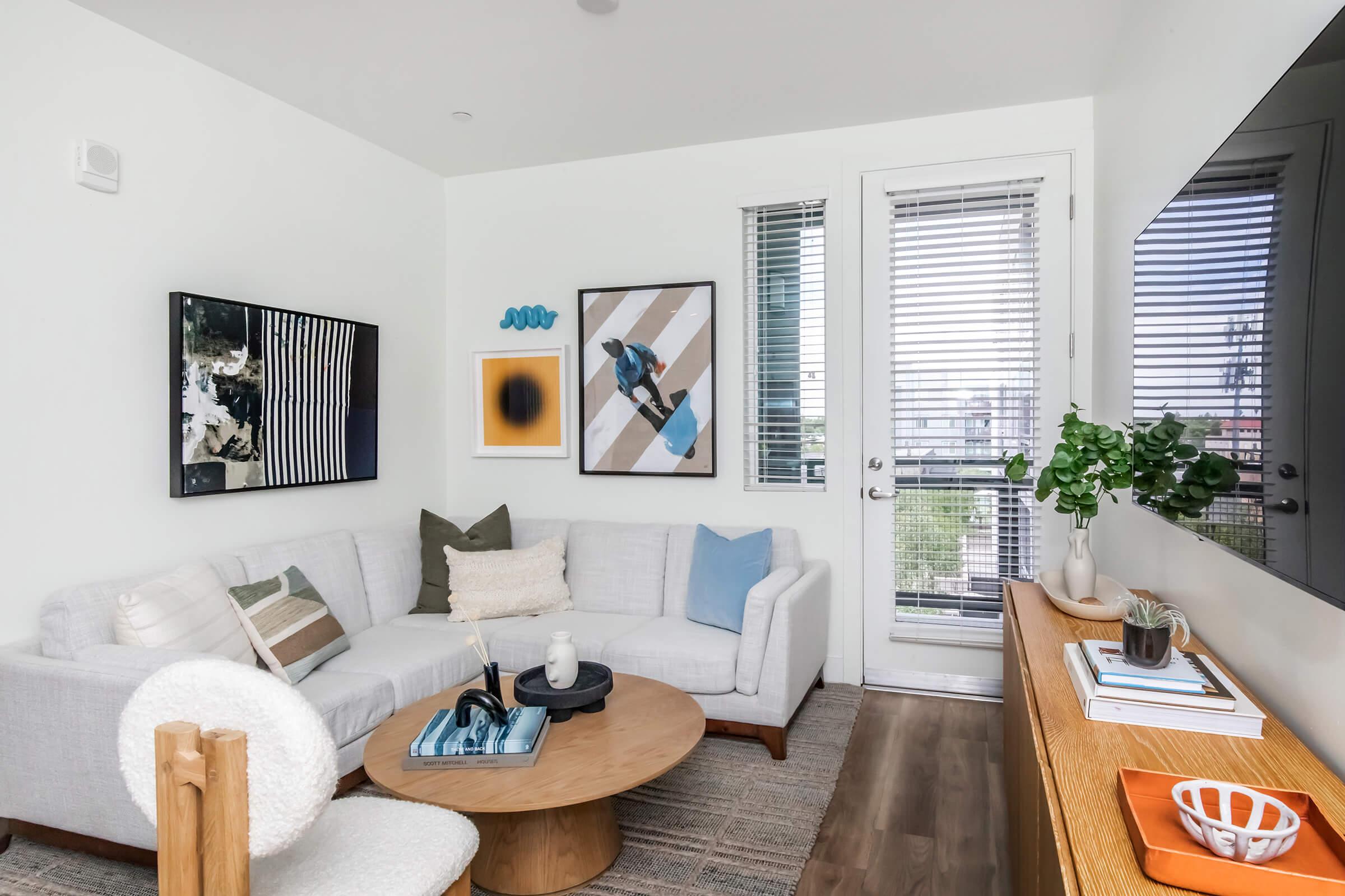 A cozy living room featuring a light-colored sectional sofa with decorative pillows, a round wooden coffee table, and a modern TV stand. The walls have contemporary art pieces, and large windows with blinds let in natural light. A green plant adds a touch of nature to the space.