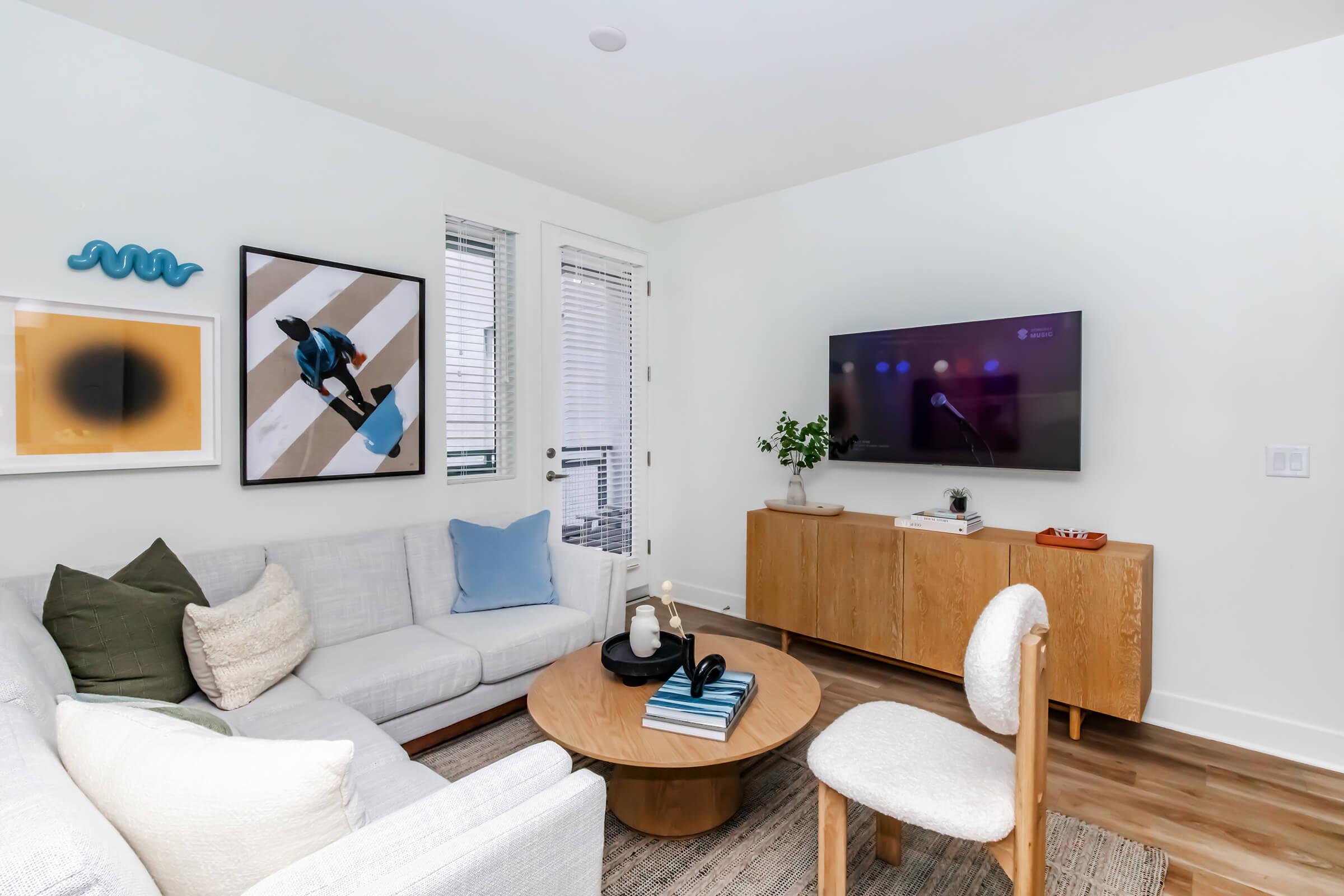 A modern living room featuring a light gray sofa with decorative pillows, a round coffee table, and a wooden sideboard with a TV mounted on the wall. The room has large windows allowing natural light, and art pieces, including a framed print and a decorative blue wall piece, enhance the decor.