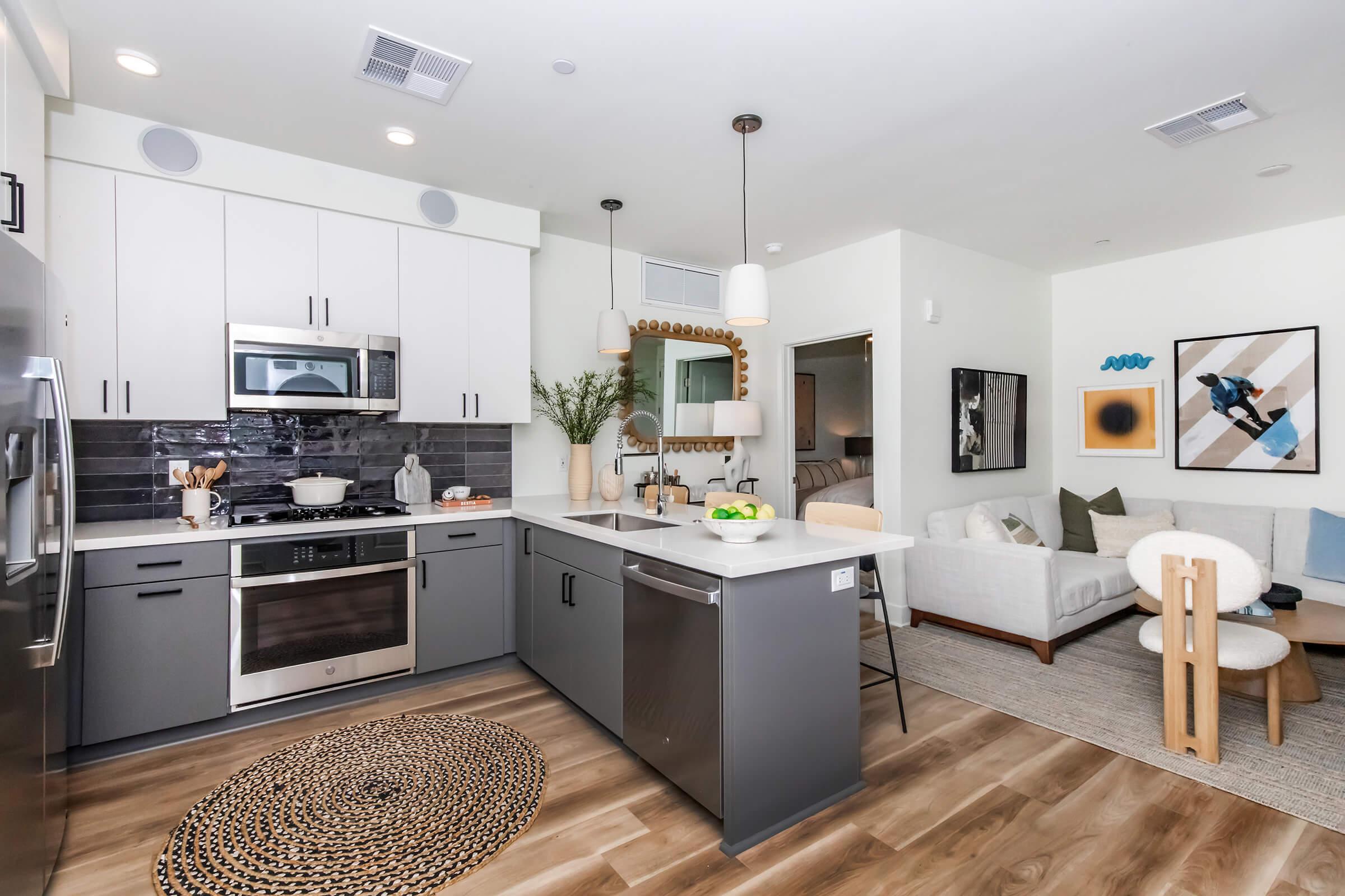 Modern kitchen featuring stainless steel appliances, sleek gray cabinets, and a central island with bar seating. The open-concept layout includes a cozy living area with a light-colored sofa, decorative artwork, and a round woven rug on the floor, creating a stylish and inviting atmosphere.