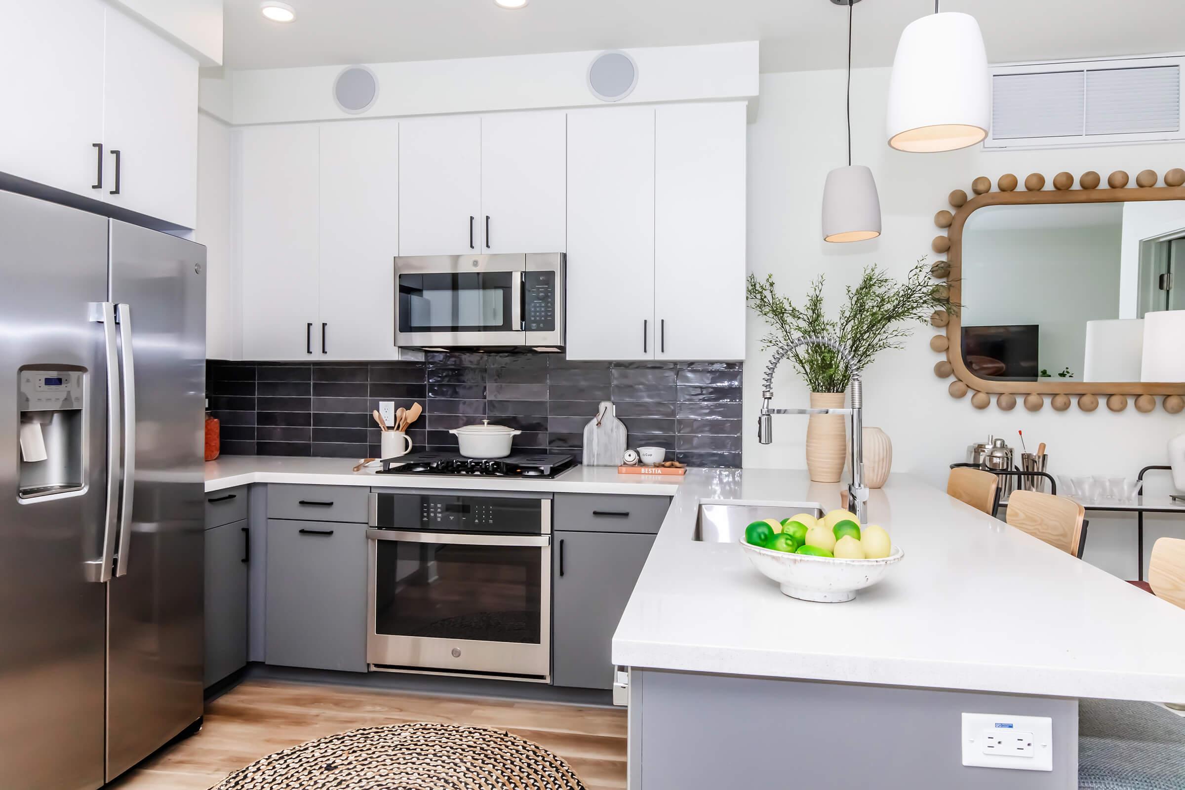 Modern kitchen featuring stainless steel appliances, including a refrigerator and microwave. The white cabinetry contrasts with dark tile backsplash. A bowl of green apples sits on a light-colored countertop, and a round rug adds texture to the floor. Warm lighting enhances the inviting atmosphere.