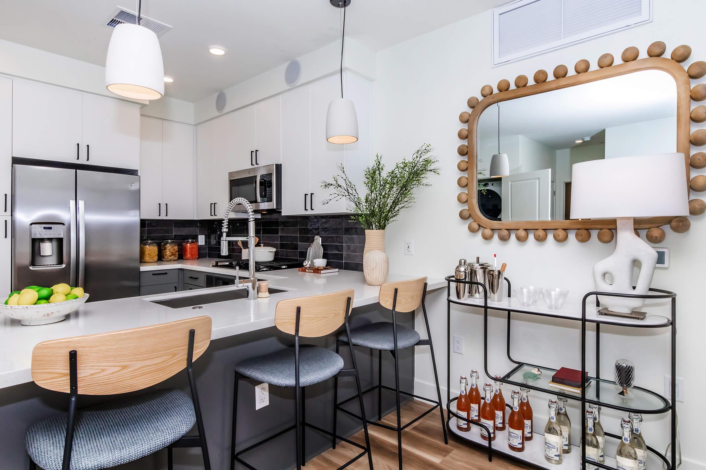 Modern kitchen with a sleek design featuring stainless steel appliances, white cabinetry, and a dark tiled backsplash. A large mirror with a decorative frame hangs above a countertop with bar stools. Fresh fruit is displayed in a bowl, and a drink cart is neatly arranged with bottles and glassware.