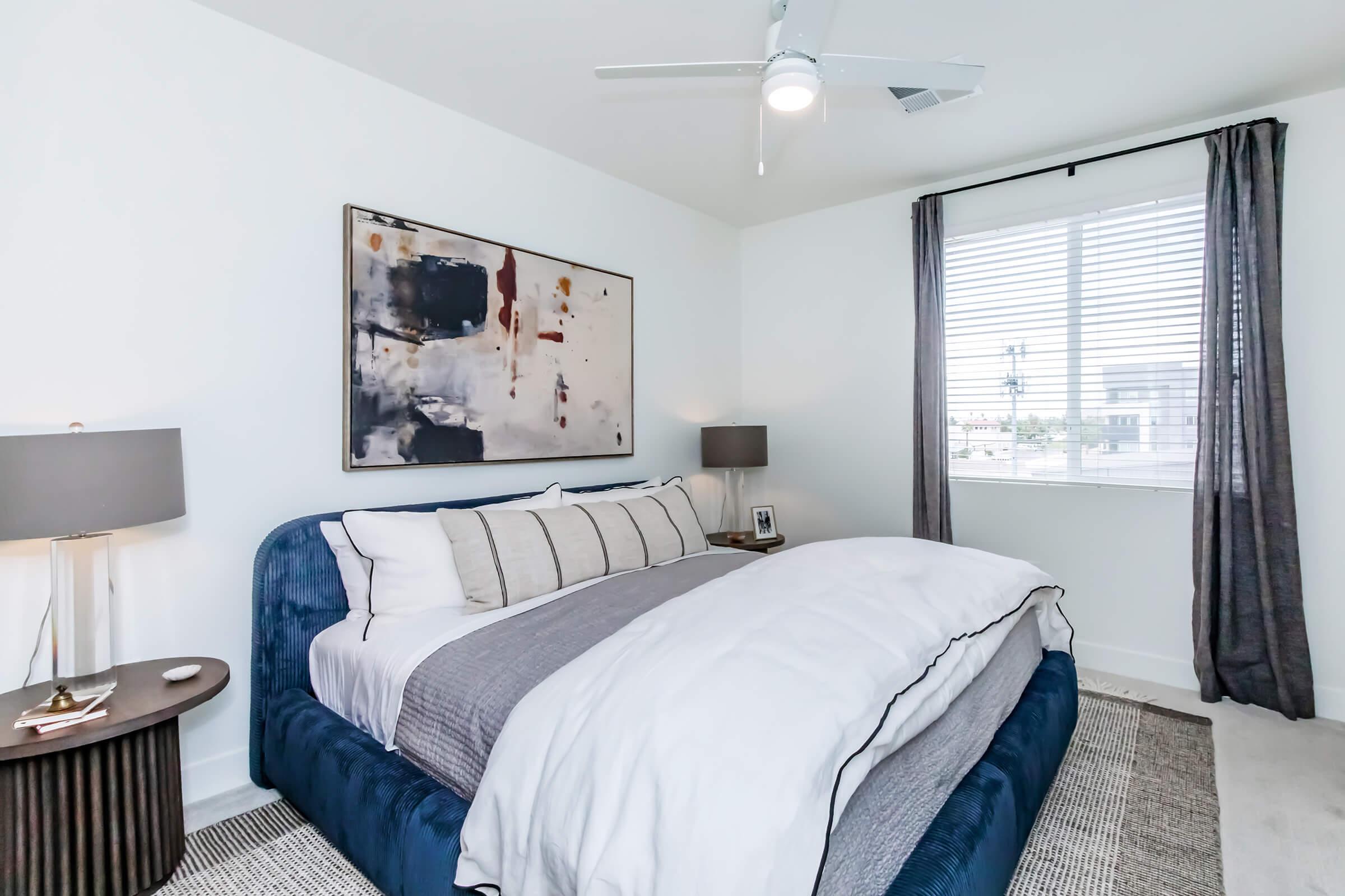 A modern bedroom featuring a stylish blue bed with white and gray bedding. Two bedside lamps flank the bed on either side. A large abstract painting hangs above the bed, and a window with blinds allows natural light to enter, complemented by dark curtains. A round table sits next to the bed.