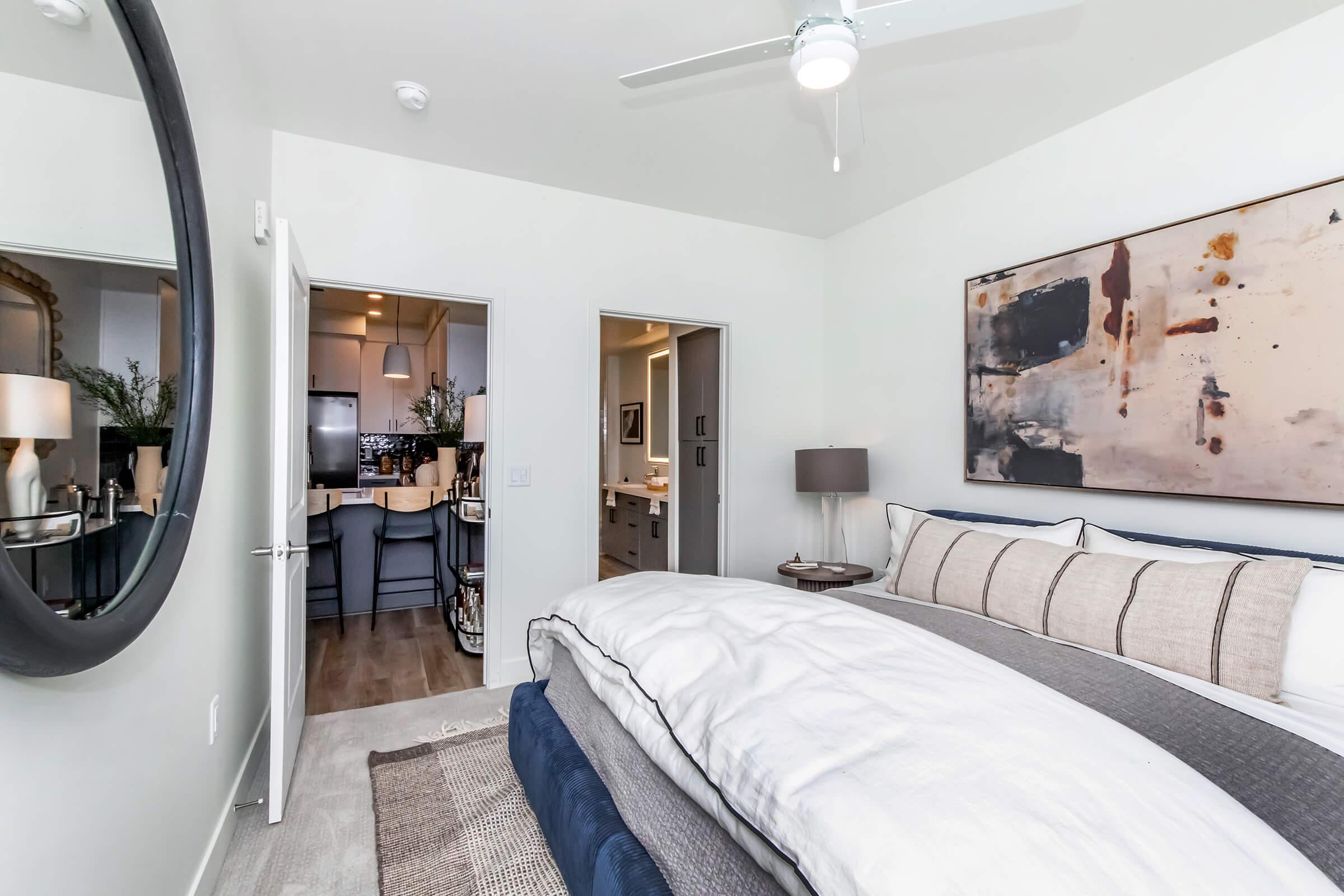 A well-decorated bedroom featuring a large bed with multiple pillows and a cozy blanket. A round mirror hangs on the wall next to the bed. In the background, there's an open doorway leading to a kitchen and a bathroom. The room has neutral colors, modern decor, and a ceiling fan.
