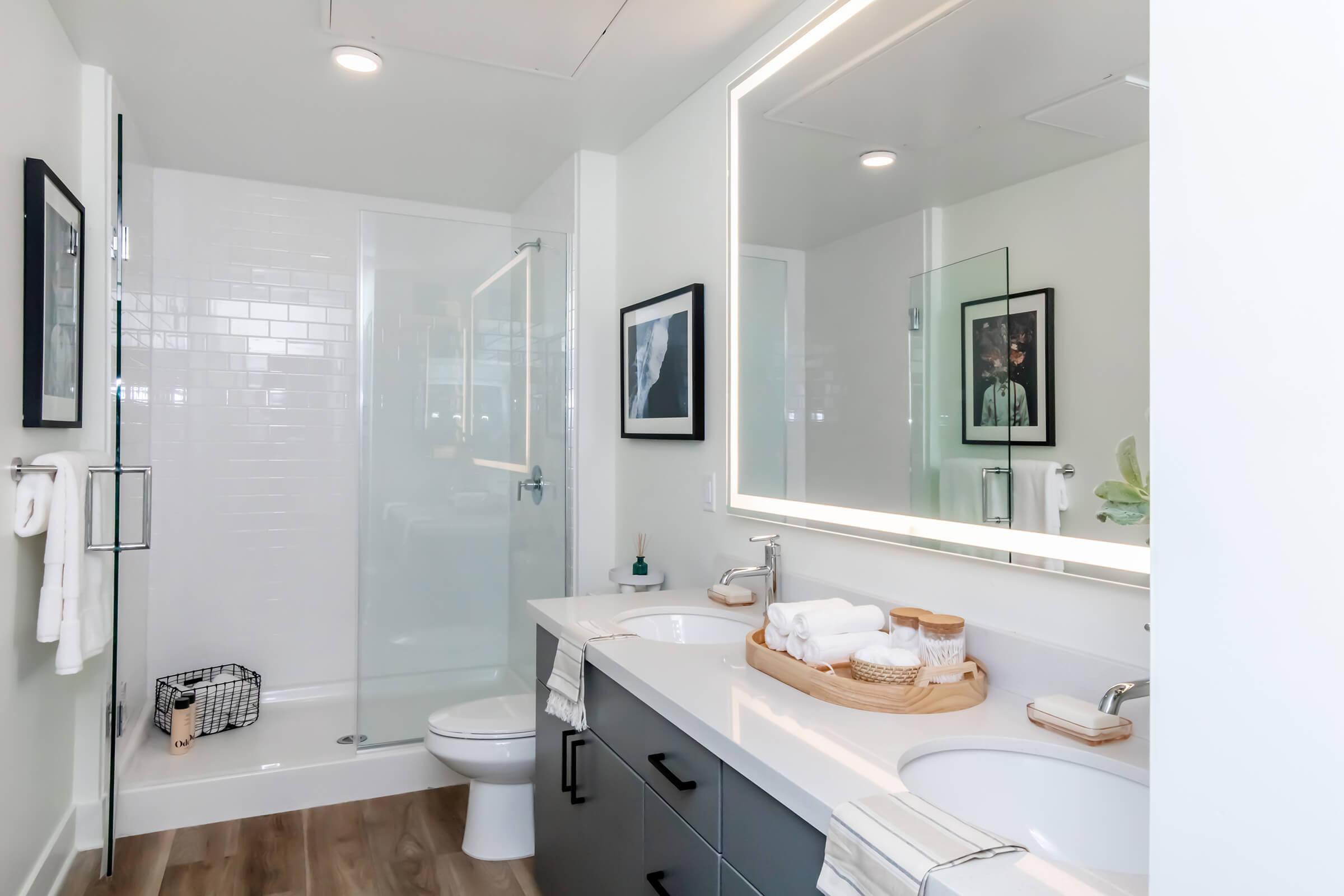 Modern bathroom featuring a glass shower, a double vanity with white countertops, and a large illuminated mirror. Decor includes a black-and-white framed photo on the wall and neatly arranged towels and toiletries on the countertop. The space has light-colored walls and wooden flooring.