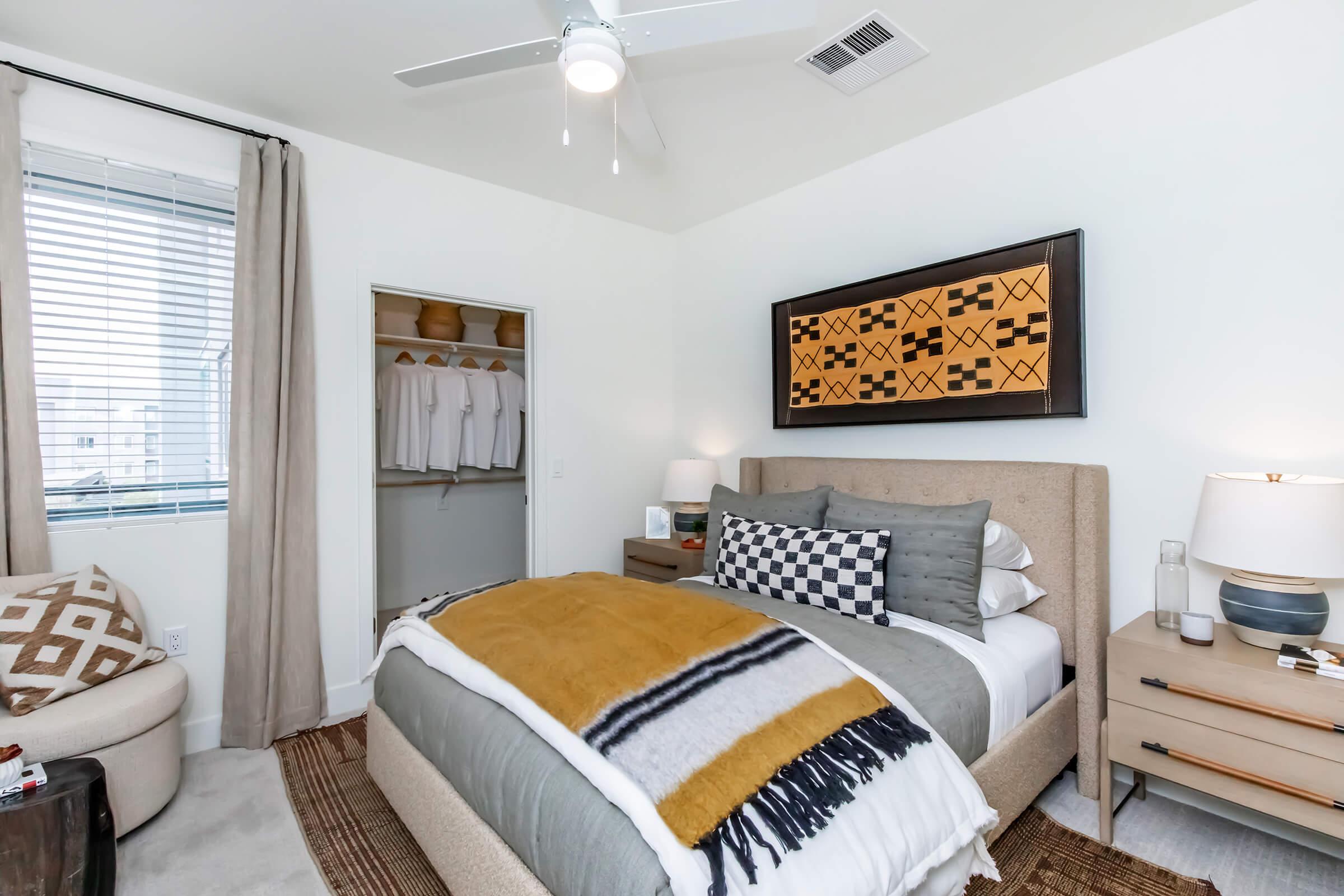 A cozy bedroom featuring a neutral color palette. The room includes a bed with a patterned blanket and decorative pillows, a nightstand with a lamp and decorative items, a plush chair, and a small closet with neatly arranged clothing. Natural light streams in from a window with light curtains.
