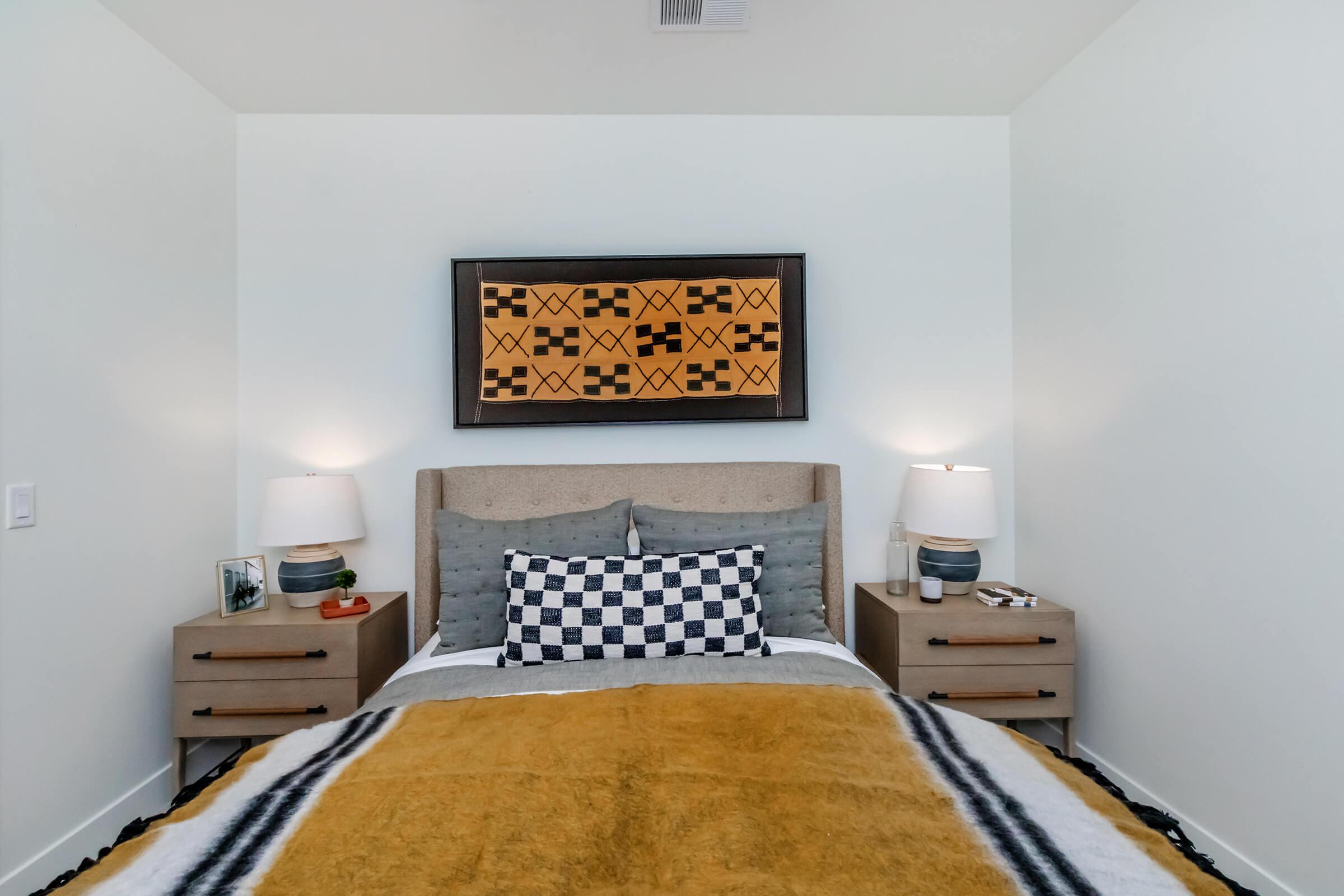 A cozy bedroom featuring a queen-sized bed with a mustard yellow throw blanket and a patterned gray and white pillow. There are two wooden nightstands with lamps on each side of the bed. A decorative artwork is hung above the headboard, and the walls are painted white, creating a bright and inviting atmosphere.