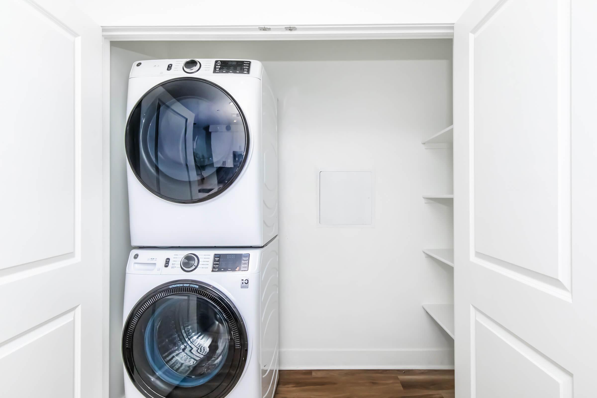 A modern laundry closet featuring a stacked white washer and dryer. The appliances have large glass doors, and the space is minimalistic with white walls and a wooden floor. There are empty shelves on the right side, providing additional storage options.