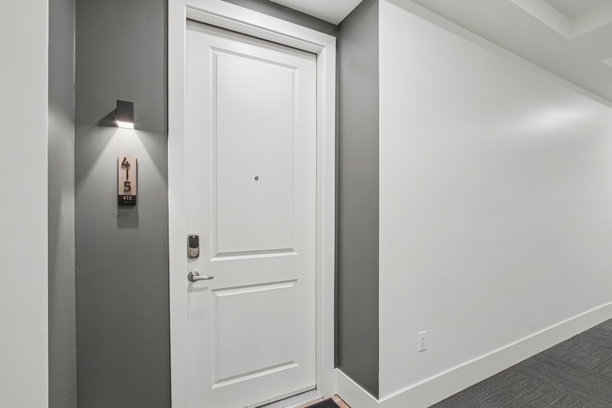 A white door with a small round peephole, located in a modern hallway. The door is numbered 415 and includes a sleek silver handle. A wall-mounted light illuminates the area next to the door, which is set against a contrasting gray wall. The flooring is a textured material, enhancing the contemporary feel of the space.