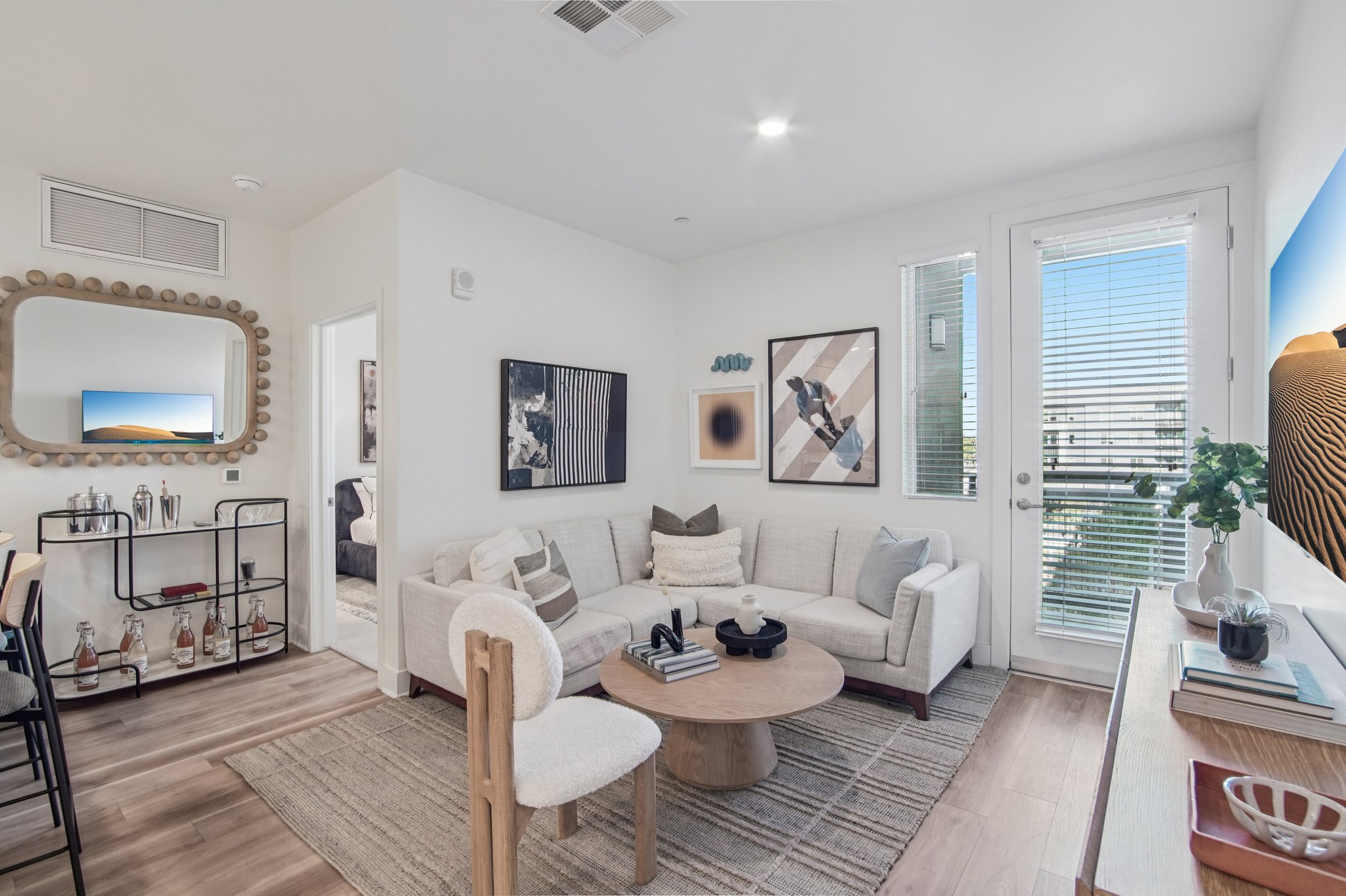 A modern living room featuring a white sectional sofa with decorative pillows, a round wooden coffee table, and a console table with plants. The walls are adorned with framed artworks. A large window allows natural light, and a mirror adds depth to the space. A dining area with black chairs is visible in the background.