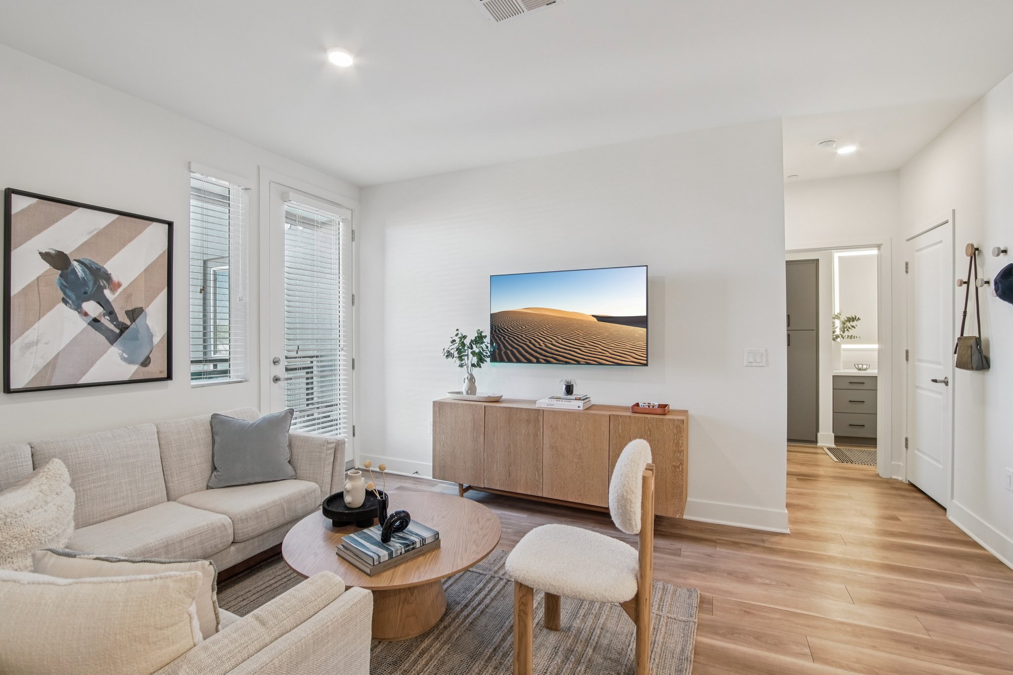 A modern living room featuring a light-colored couch with cushions, a round coffee table, and a wooden TV unit displaying a desert landscape on the screen. Natural light enters through large windows, and a small plant adds a touch of greenery. A bathroom is visible through a door in the background, enhancing the contemporary design.