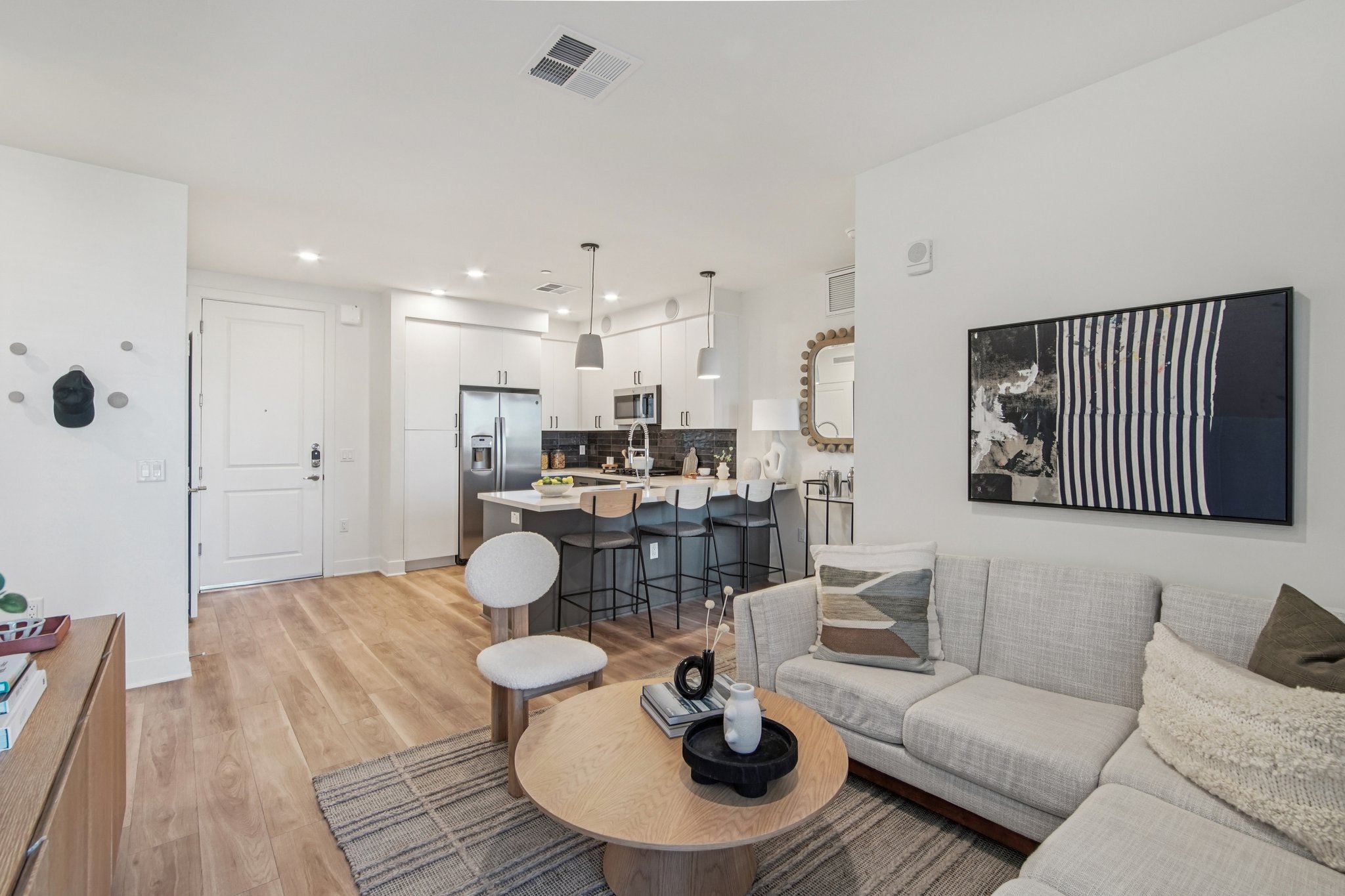 Modern living room and kitchen area featuring a light-colored sectional sofa, a round coffee table, and a dining space with bar stools. The kitchen has stainless steel appliances and a large island. Light wood flooring complements the neutral decor, and artwork is displayed on the wall.