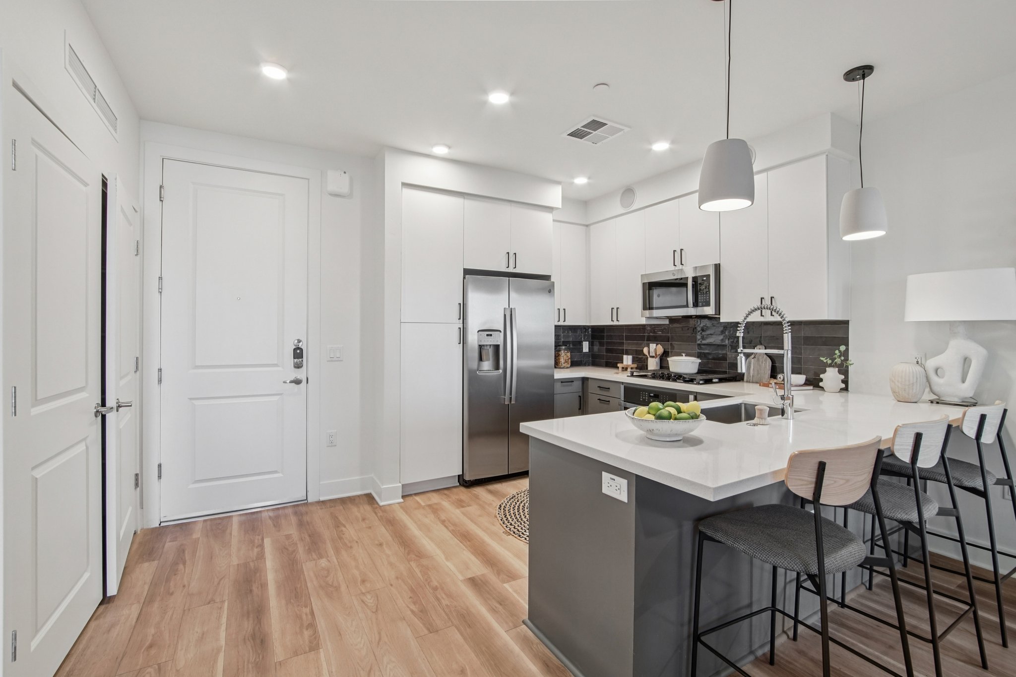 Modern kitchen featuring sleek white cabinets, stainless steel appliances, and a dark tiled backsplash. A large island with bar stools is in the foreground, and the space is illuminated by pendant lights. Natural wood flooring adds warmth to the contemporary design.