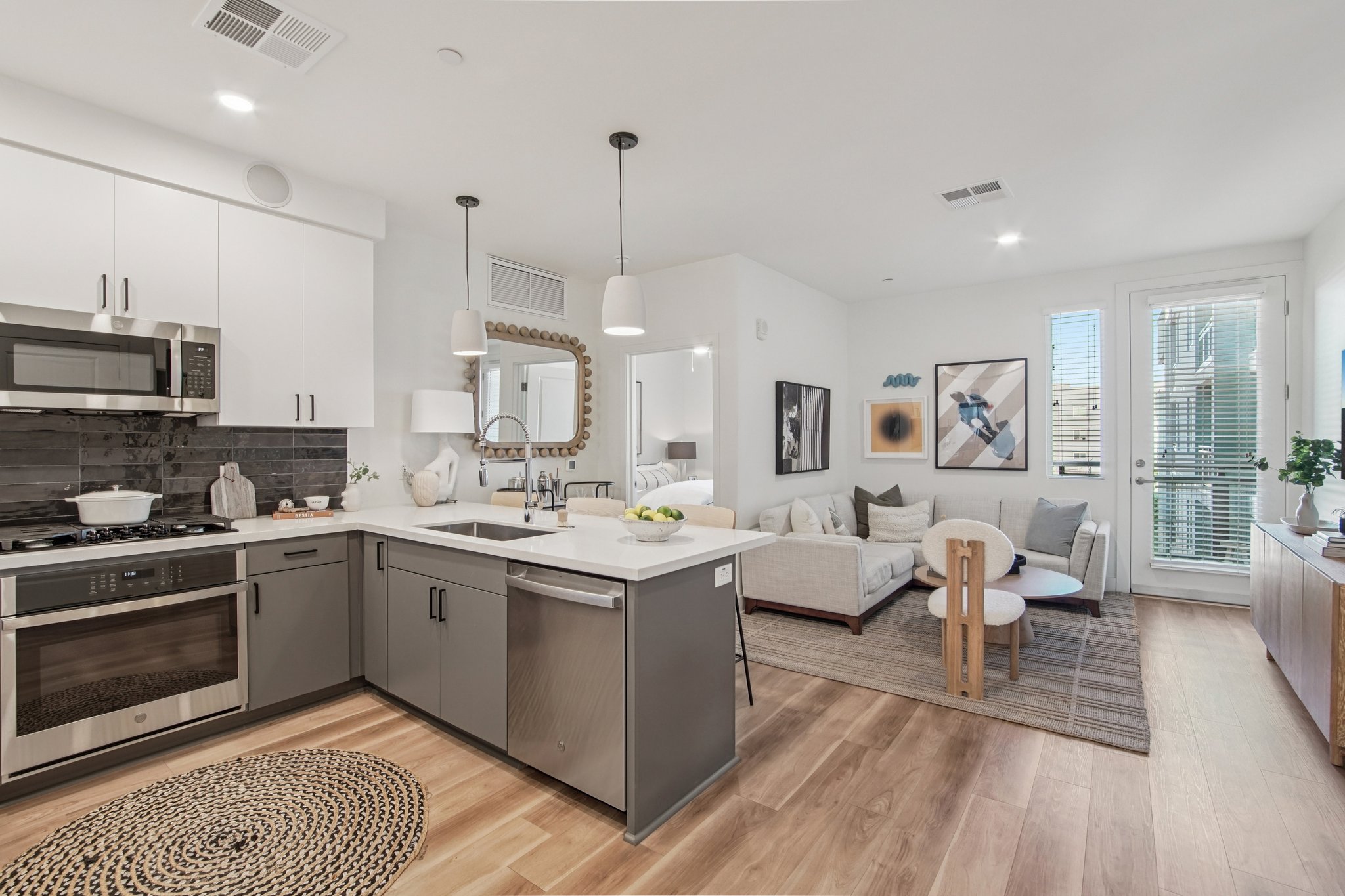 Modern kitchen and living room in an open-concept apartment. The kitchen features sleek gray cabinetry, stainless steel appliances, and a center island with a breakfast bar. The living area includes a light-colored sofa, a rug, and large windows allowing natural light. Decorative artwork and plants add warmth to the space.