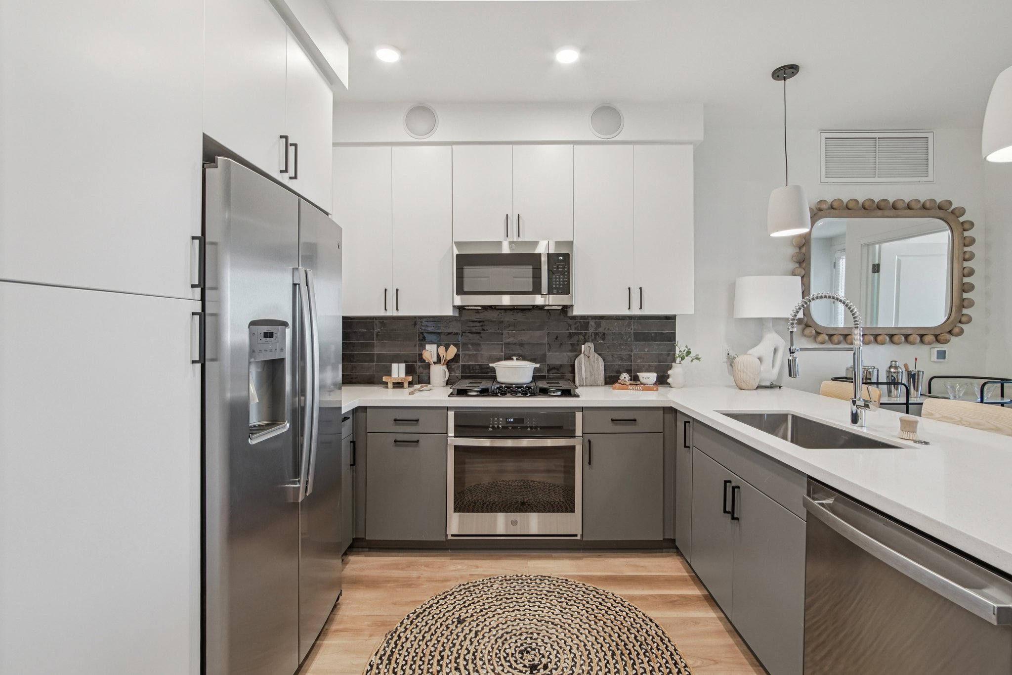A modern kitchen featuring stainless steel appliances, including a refrigerator and oven. The cabinets are a mix of white and gray, with a sleek countertop. A round, woven rug is on the floor, and there are small decorative items on the counter. The space is bright and well-lit, with contemporary fixtures.