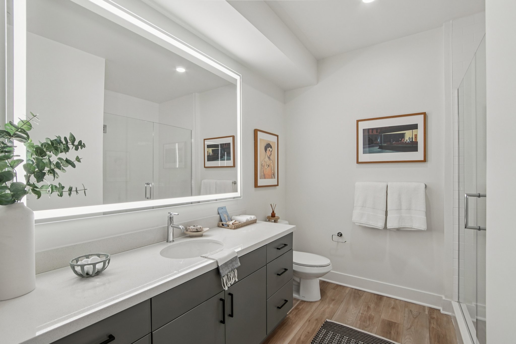 A modern bathroom featuring a large illuminated mirror above a white countertop sink. The space includes dark cabinetry, a framed artwork on the wall, neatly arranged towels, and a shower area with glass doors. A plant adds a touch of greenery to the serene, minimalist design.