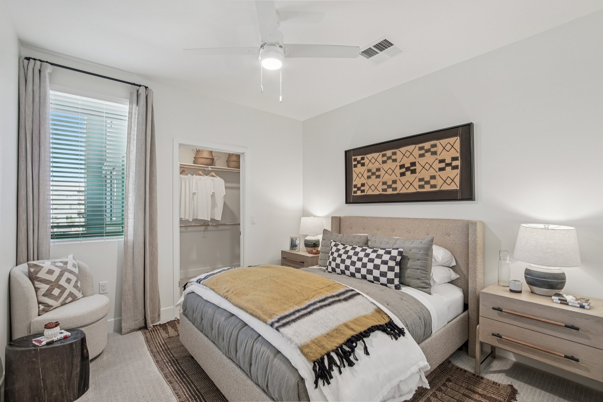 A cozy bedroom featuring a queen-sized bed with decorative pillows and a soft throw blanket. There are two bedside tables with lamps, a small armchair, and a closet with hanging clothes. Natural light filters through the window, and the walls are adorned with a patterned artwork. A ceiling fan adds to the comfort.