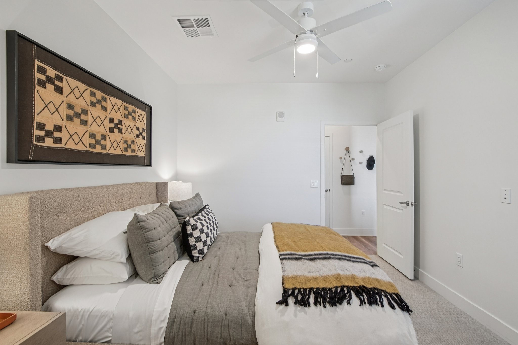 A well-decorated bedroom featuring a beige upholstered bed with gray and plaid pillows, a neutral throw blanket draped across the foot of the bed, and an artistic wall hanging above. A ceiling fan provides ventilation, and a door leads to another room. The walls are painted in light colors, creating a tranquil atmosphere.