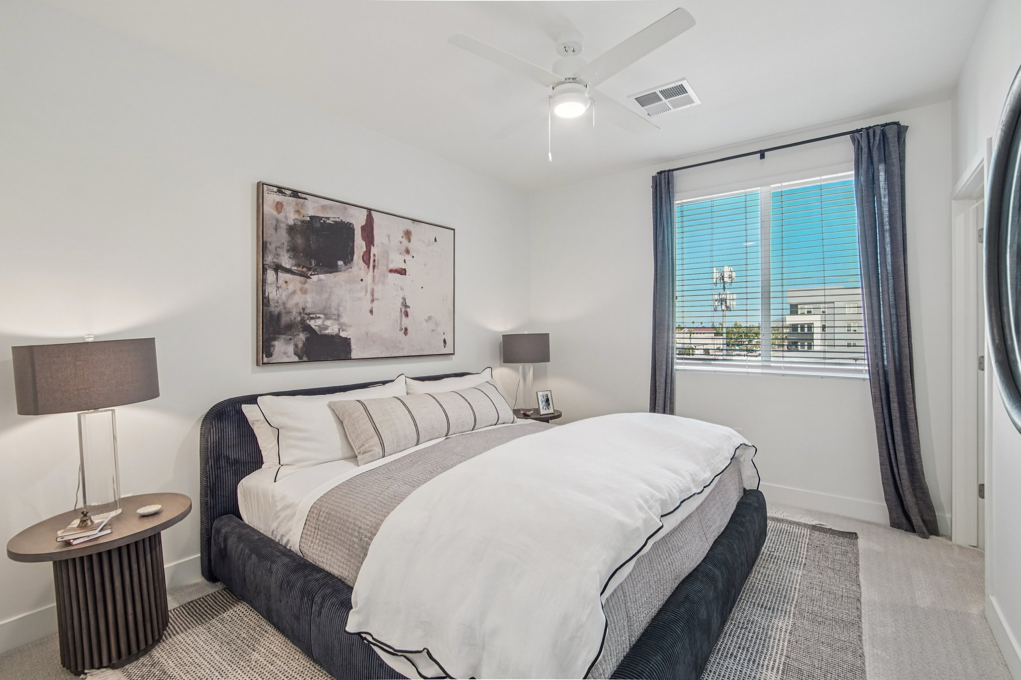 A modern bedroom featuring a large bed with white and gray bedding, a dark upholstered headboard, and two bedside lamps. There's a circular wall mirror, textured rug, and a window with sheer curtains, allowing natural light to illuminate the space. Artwork hangs above the bed, adding a touch of decor.