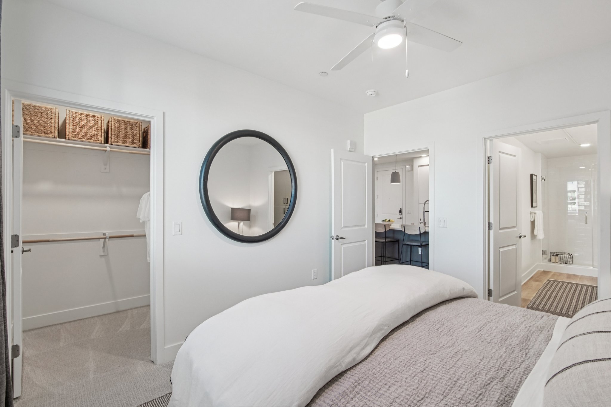A modern bedroom featuring a large bed with white linens, a round mirror on the wall, and a ceiling fan. There are two doorways leading to different areas, one with a walk-in closet containing woven baskets and another leading to a bathroom. The decor is minimalist with neutral colors and soft lighting.