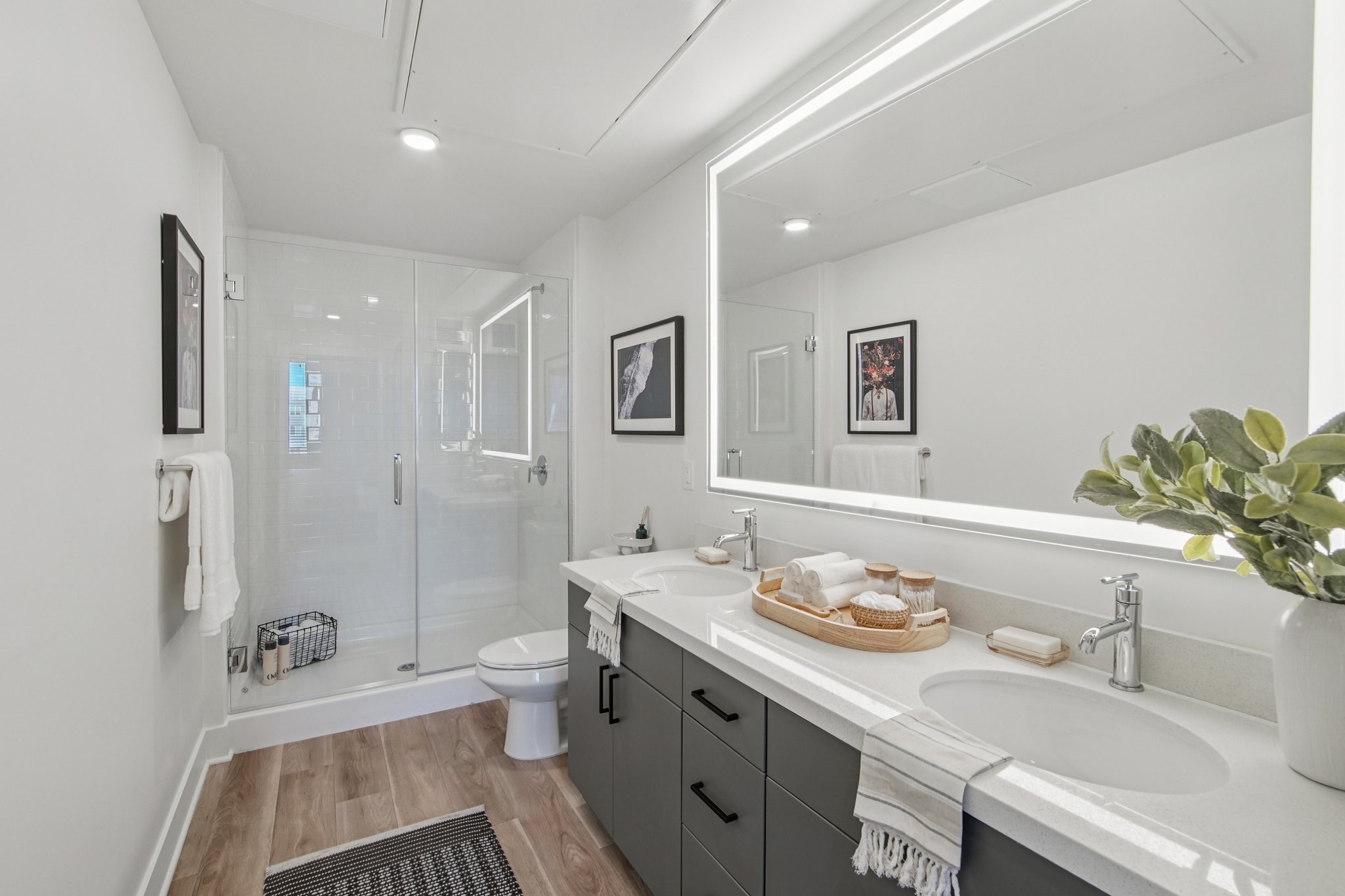 A modern bathroom featuring a double sink with a stylish vanity, a large mirror with backlighting, a walk-in shower with clear glass doors, and minimalist decor including towels and plants. The floor has light-colored wood, and framed artwork adorns the walls.