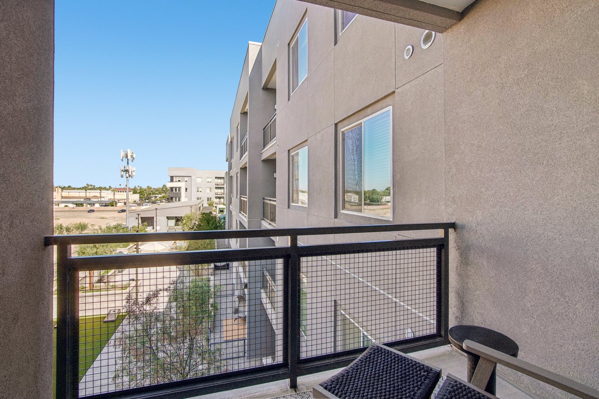 A view from a balcony showing adjacent modern buildings and a clear sky. The balcony features a railing and a small table with chairs, overlooking a landscaped area with greenery.