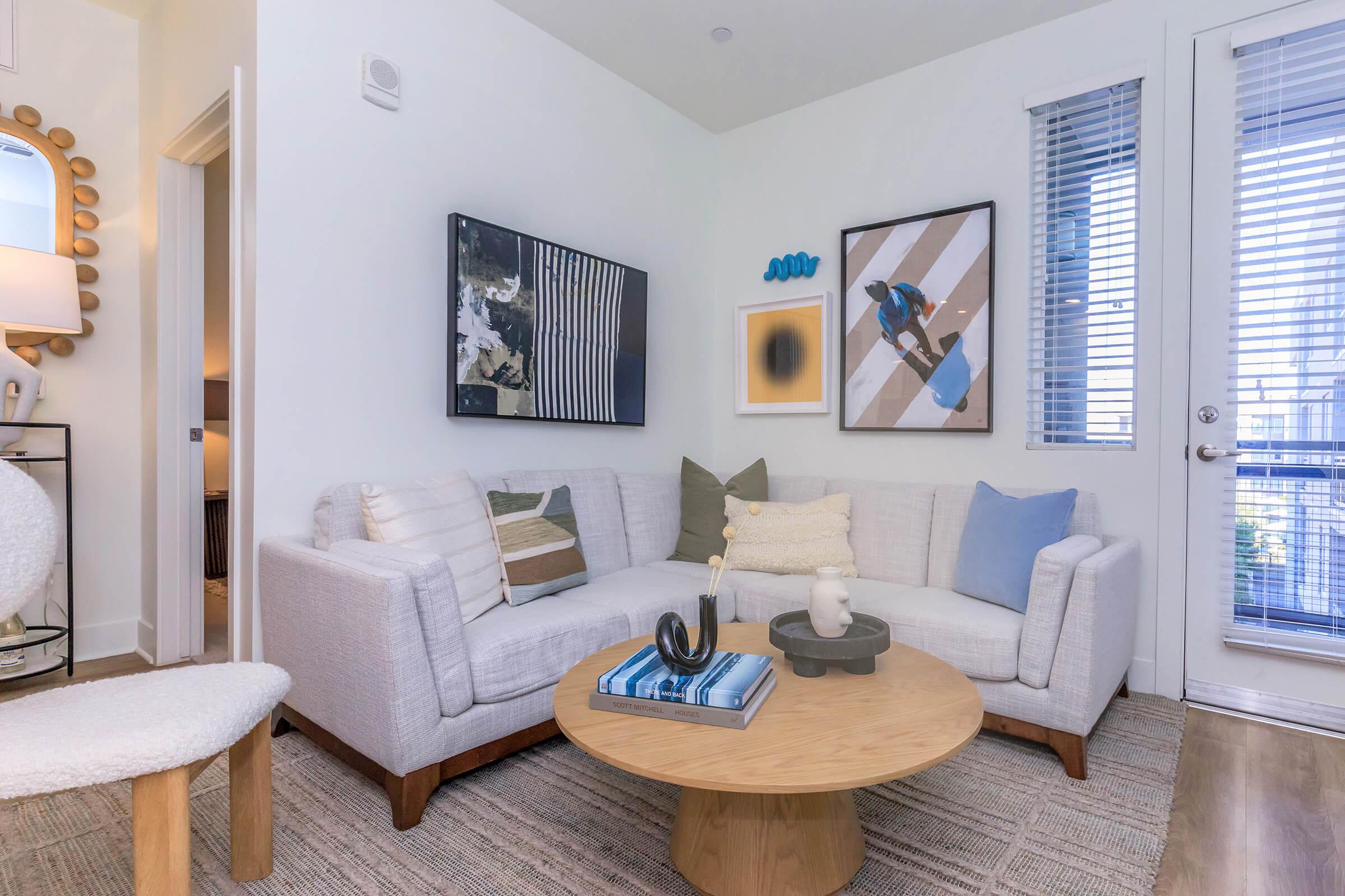A bright and modern living room featuring a light-colored sectional sofa, a round wooden coffee table, and decorative art on the walls. The space includes large windows with blinds, an accent chair, and a subtle area rug, creating a cozy and inviting atmosphere.