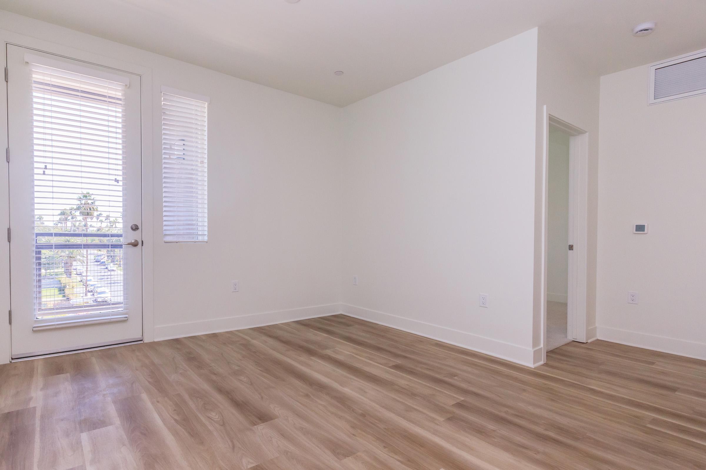 A bright, empty room with light-colored walls and wooden flooring. There are two windows with blinds on one wall, and a doorway leading to another room. The space is uncluttered and minimalistic, providing a clean, modern aesthetic.
